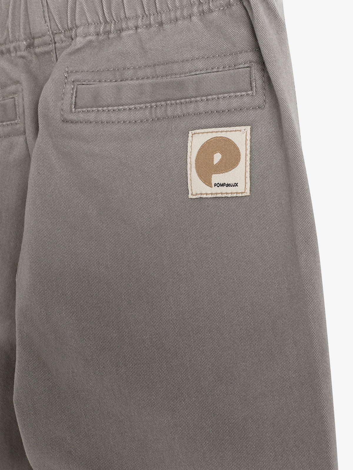 Eine graue Hose mit elastischem Bund zeigt eine Gesäßtasche und ein aufgenähtes Etikett mit dem Buchstaben „P“ und dem Text „POMPdeLUX“.