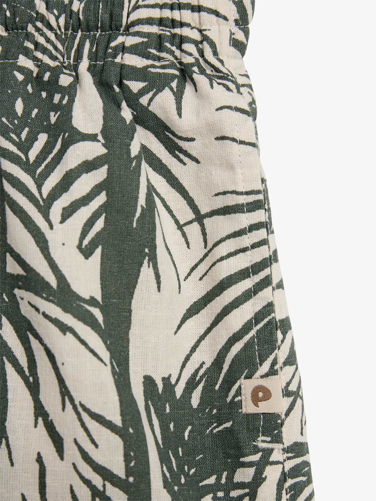 Fabric with a leafy, tropical pattern in dark green on a beige background; features an elastic waistband and a small sewn label depicting a stylized "P" in a tag.