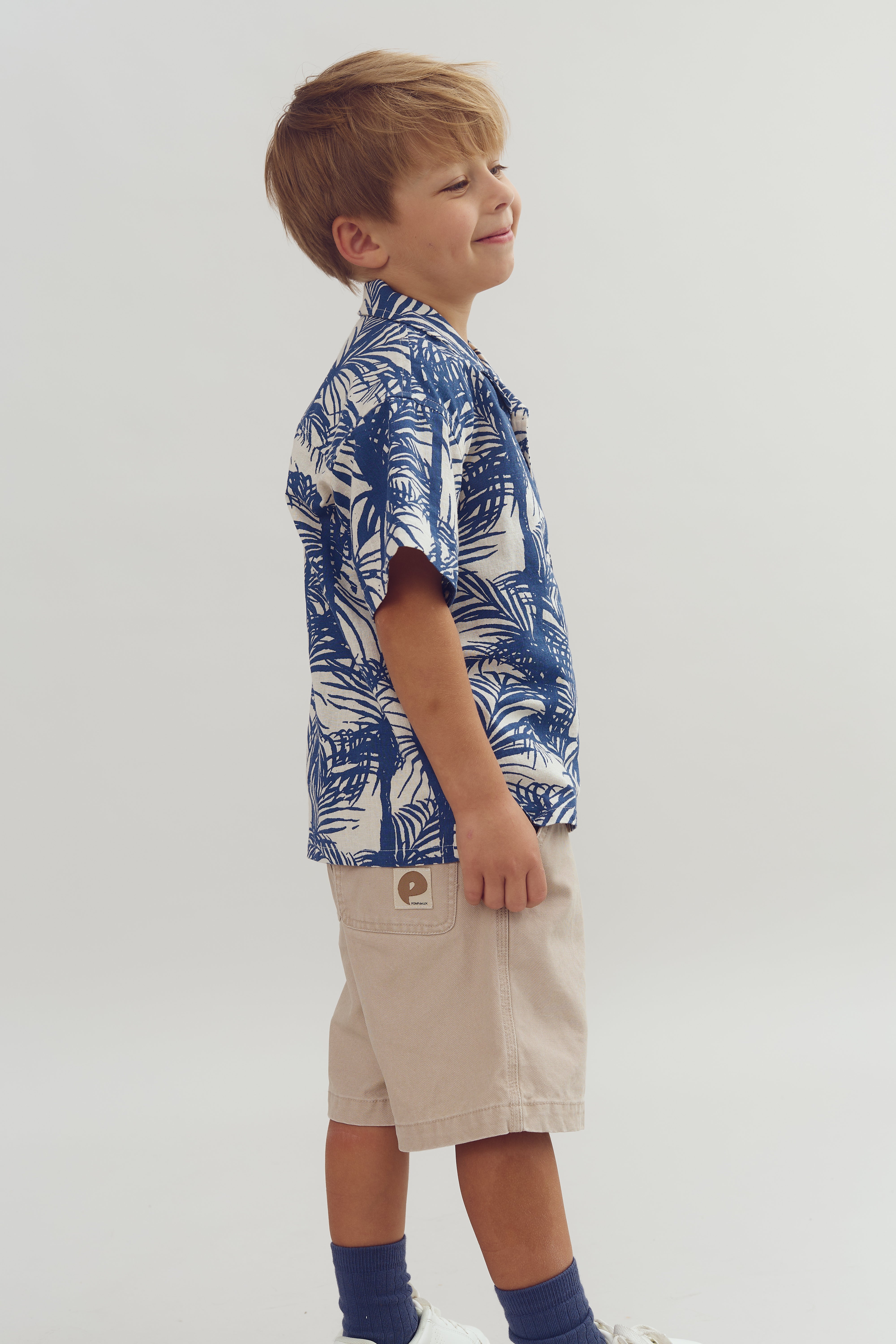 Young boy stands, smiling slightly, wearing a blue and white tropical shirt and beige shorts with a logo. He's in a bright, neutral space.