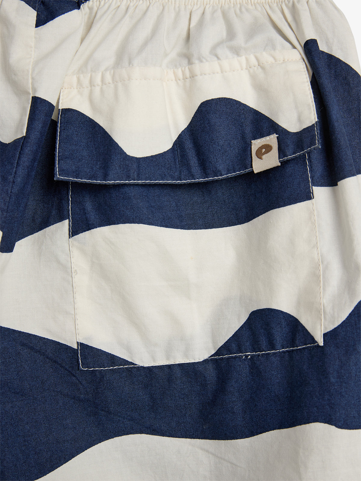 A fabric with a blue and white wave pattern features a stitched pocket with a small tag. The background is the same patterned material.