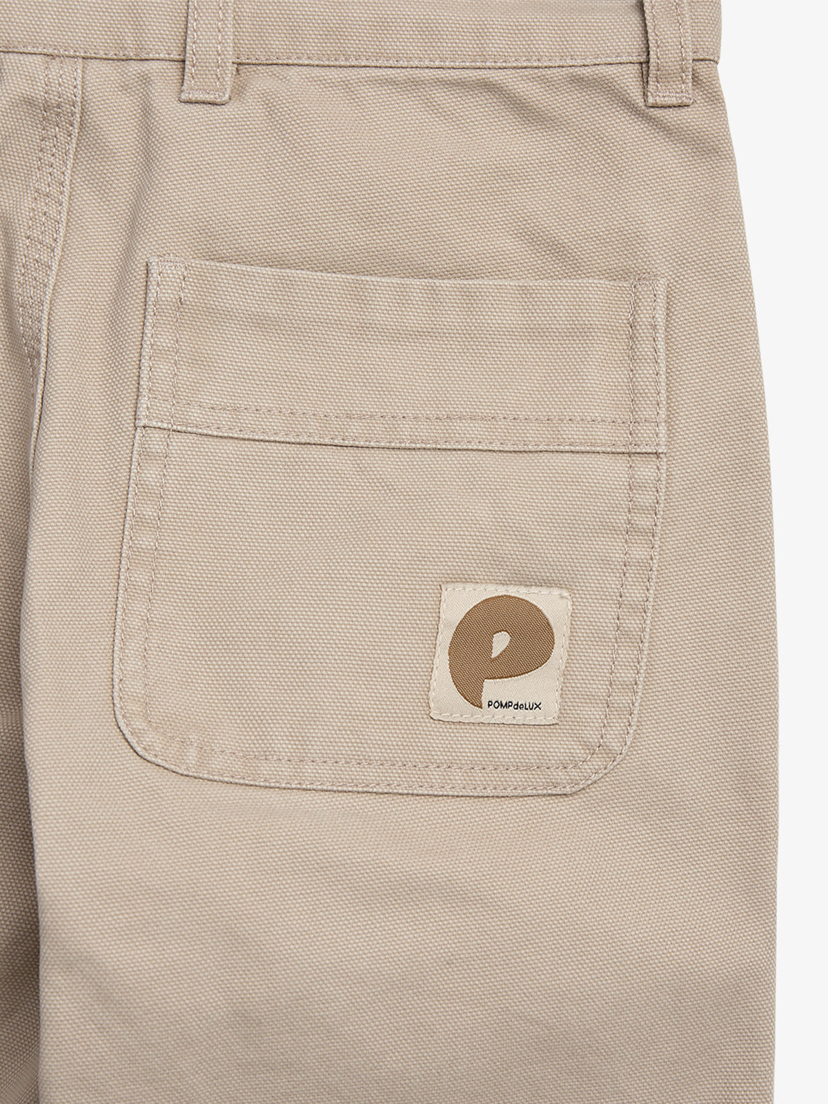 Beige pants with a rear pocket feature a square label displaying a stylized "P" and the text "POMPEIIUX." The pants are neatly stitched and have belt loops.