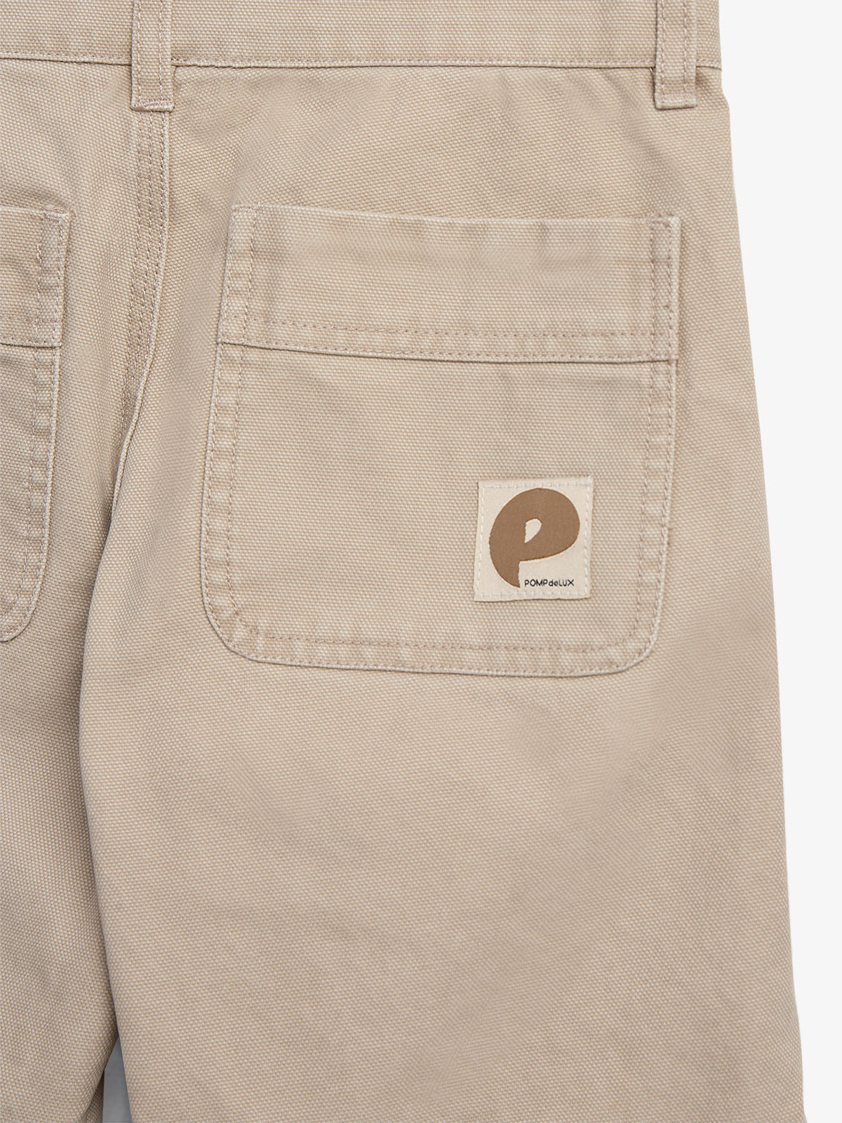 Khaki pants with visible back pocket featuring a brown "P" logo. Tag text reads "Pom Péliux." The pants are neatly stitched and positioned against a plain background.