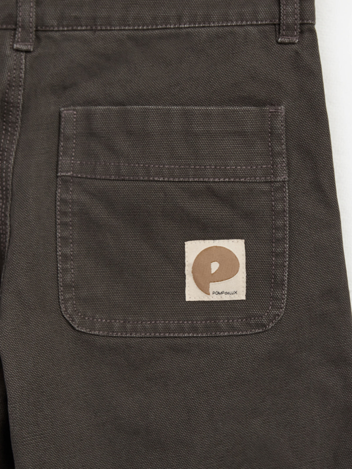 Dark fabric with a stitched pocket featuring a small white label. The label displays a beige logo resembling a lowercase "p" and the text "POMPEALUX."