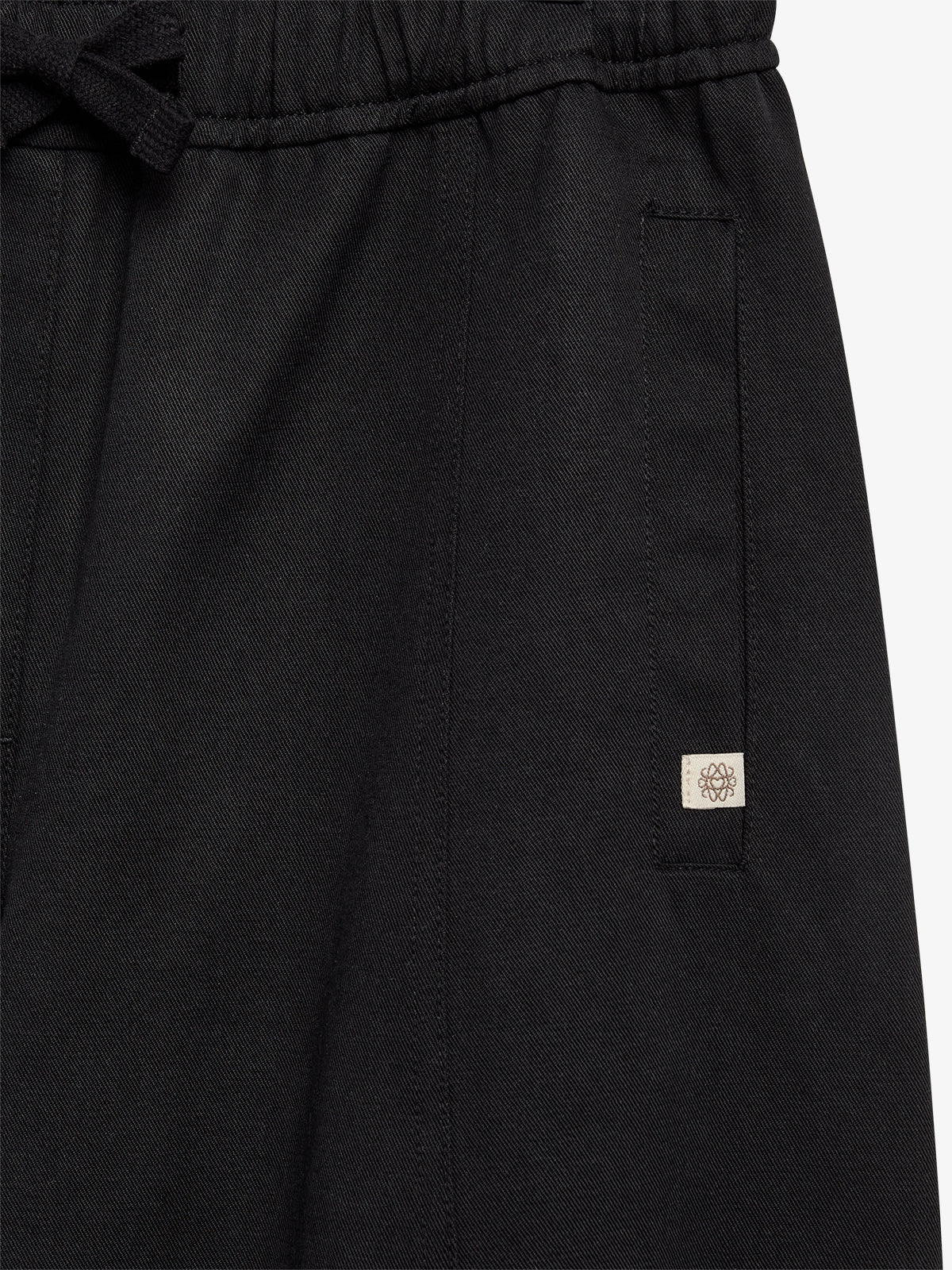 Black fabric trousers feature an elastic waistband with a drawstring, a rectangular pocket, and a small logo tag with a floral design on the pocket.