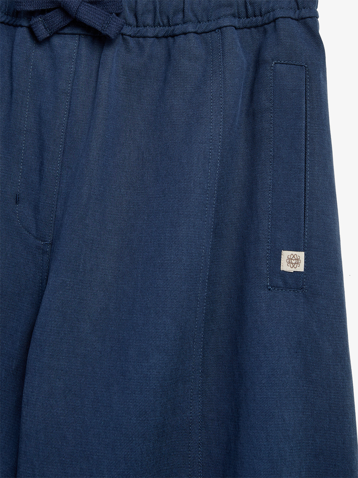 Blue pants with an elastic waistband and drawstring, featuring a seam pocket on the right. A small floral logo patch is sewn near the pocket. Background is plain white.