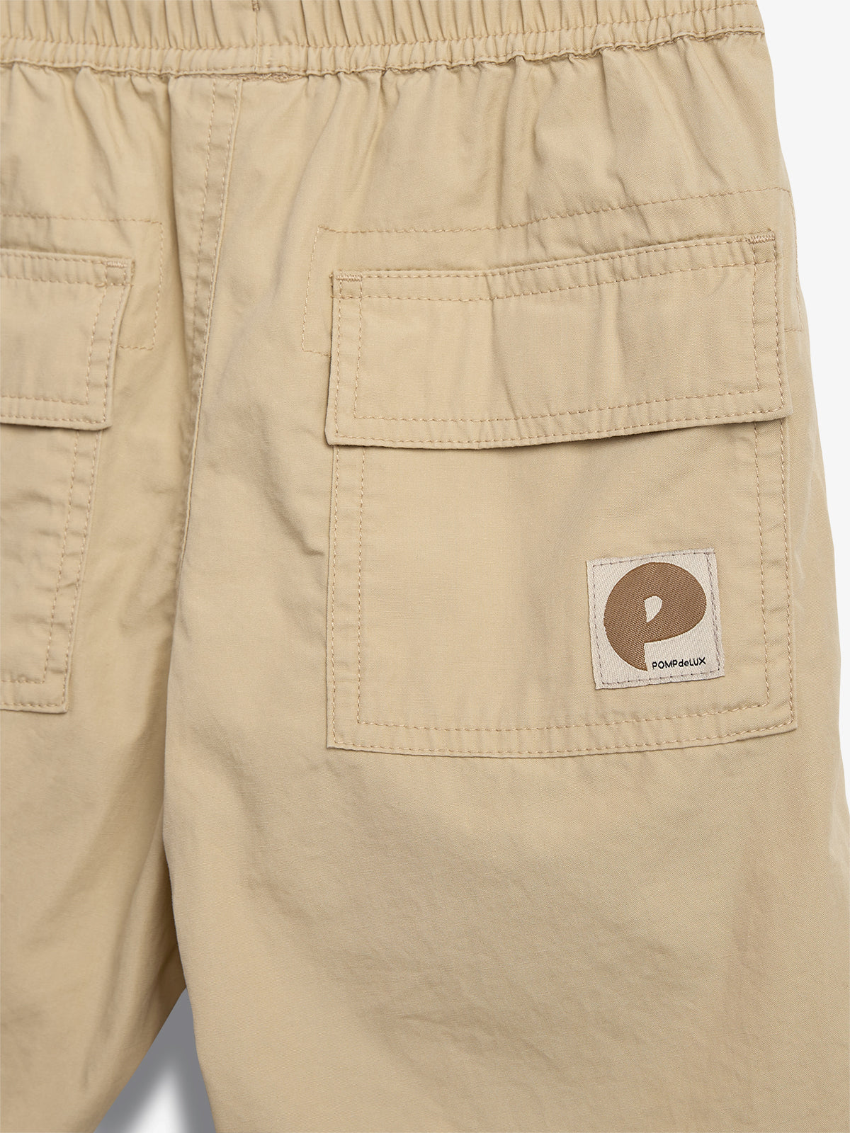 Beige shorts feature two back pockets with flaps, an elastic waistband, and a patch displaying a stylized letter "P" with the text "POMPdeLUX" on one pocket.
