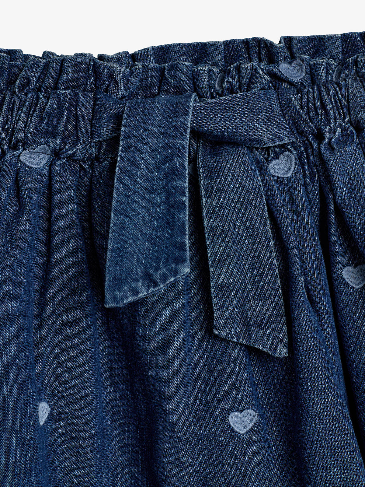 Denim fabric with an elastic waistband and a tied belt; heart patterns are subtly woven, adding texture. The material is gathered, giving a slightly ruffled appearance.