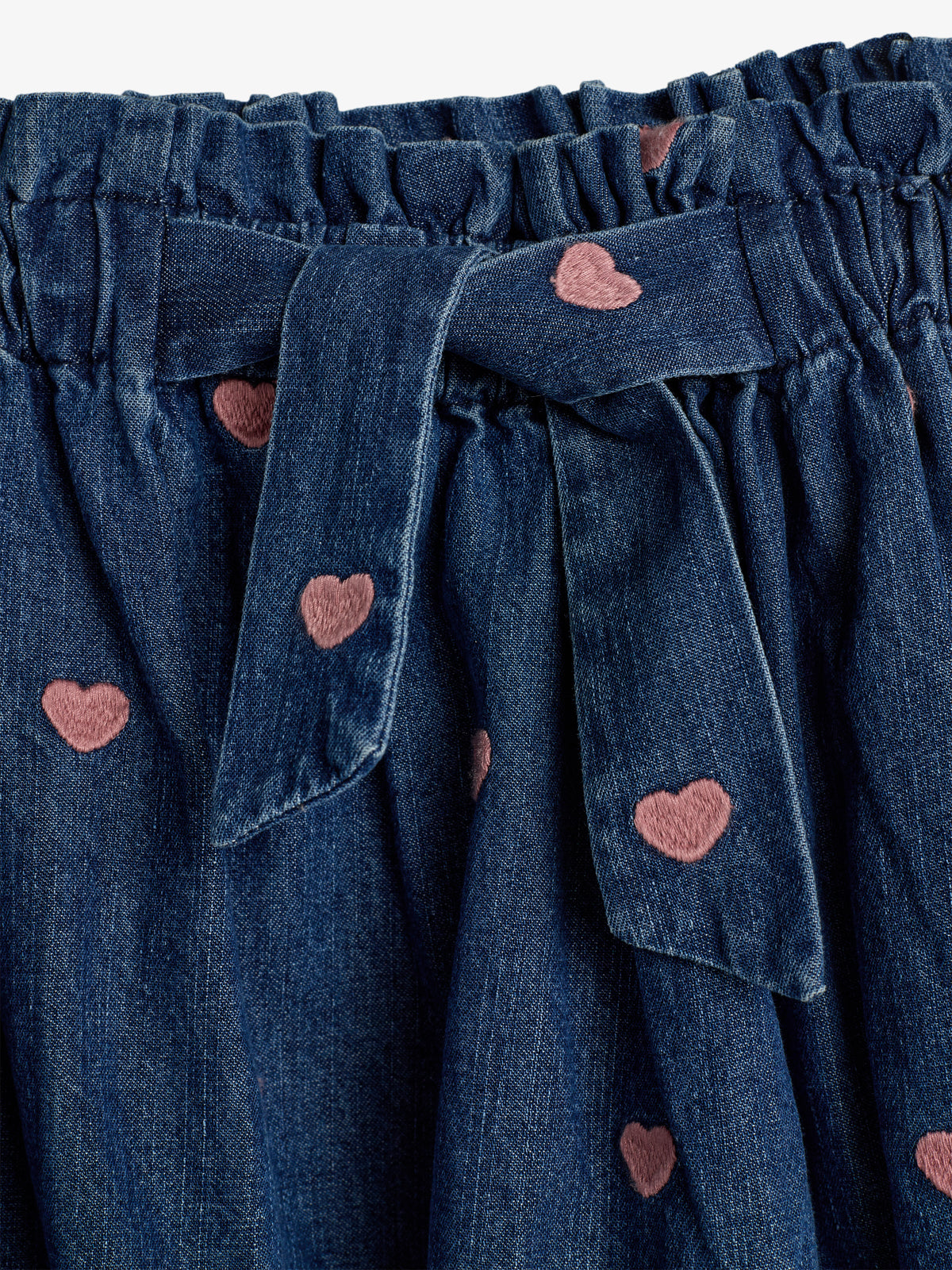 A denim fabric with a tied belt featuring pink heart patterns is gathered, creating a ruched texture, emphasizing a playful, stylish appearance.