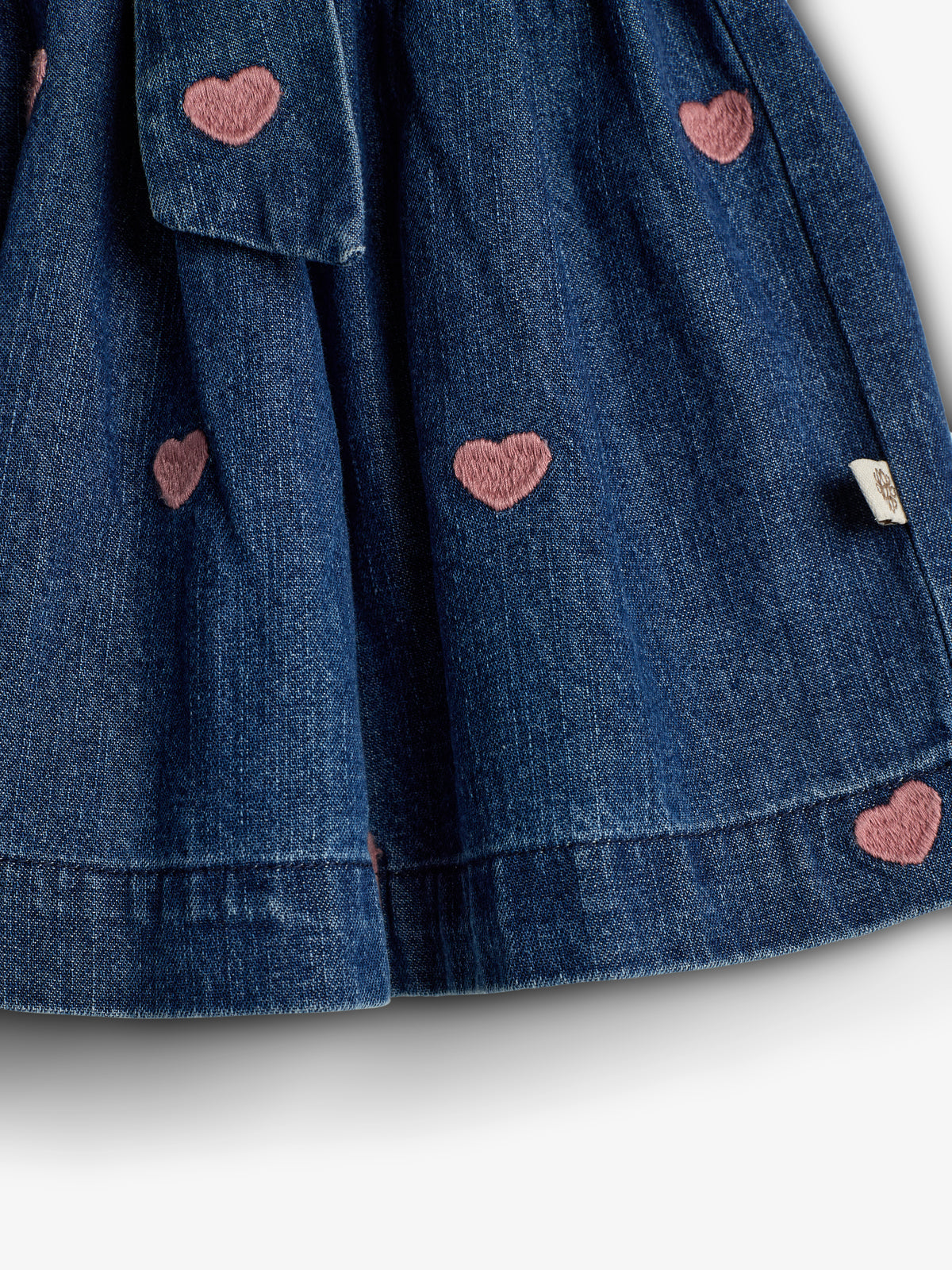 Denim fabric with pink heart embroidery patterns, forming a skirt. The tag appears on the right. The skirt folds create a textured appearance against a plain white background.