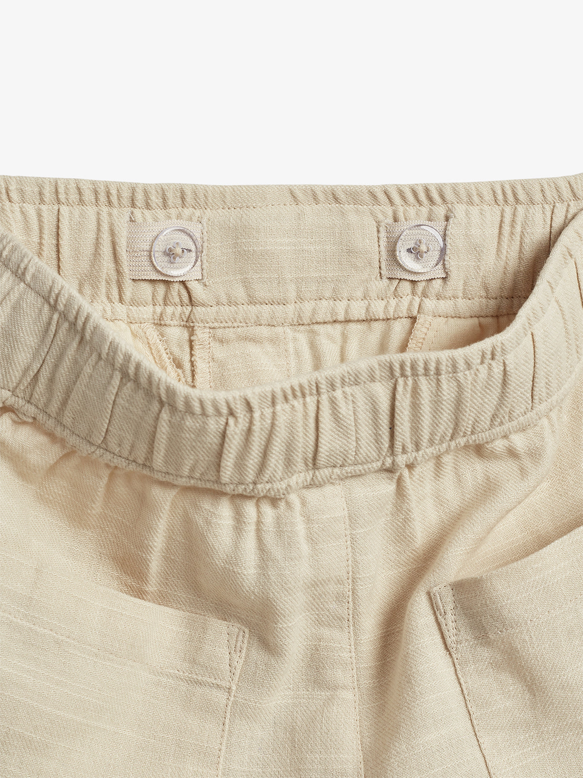 Beige elastic waistband pants feature button adjustments inside the waistband for fit, with visible stitching and pockets, placed in a neutral indoor environment.