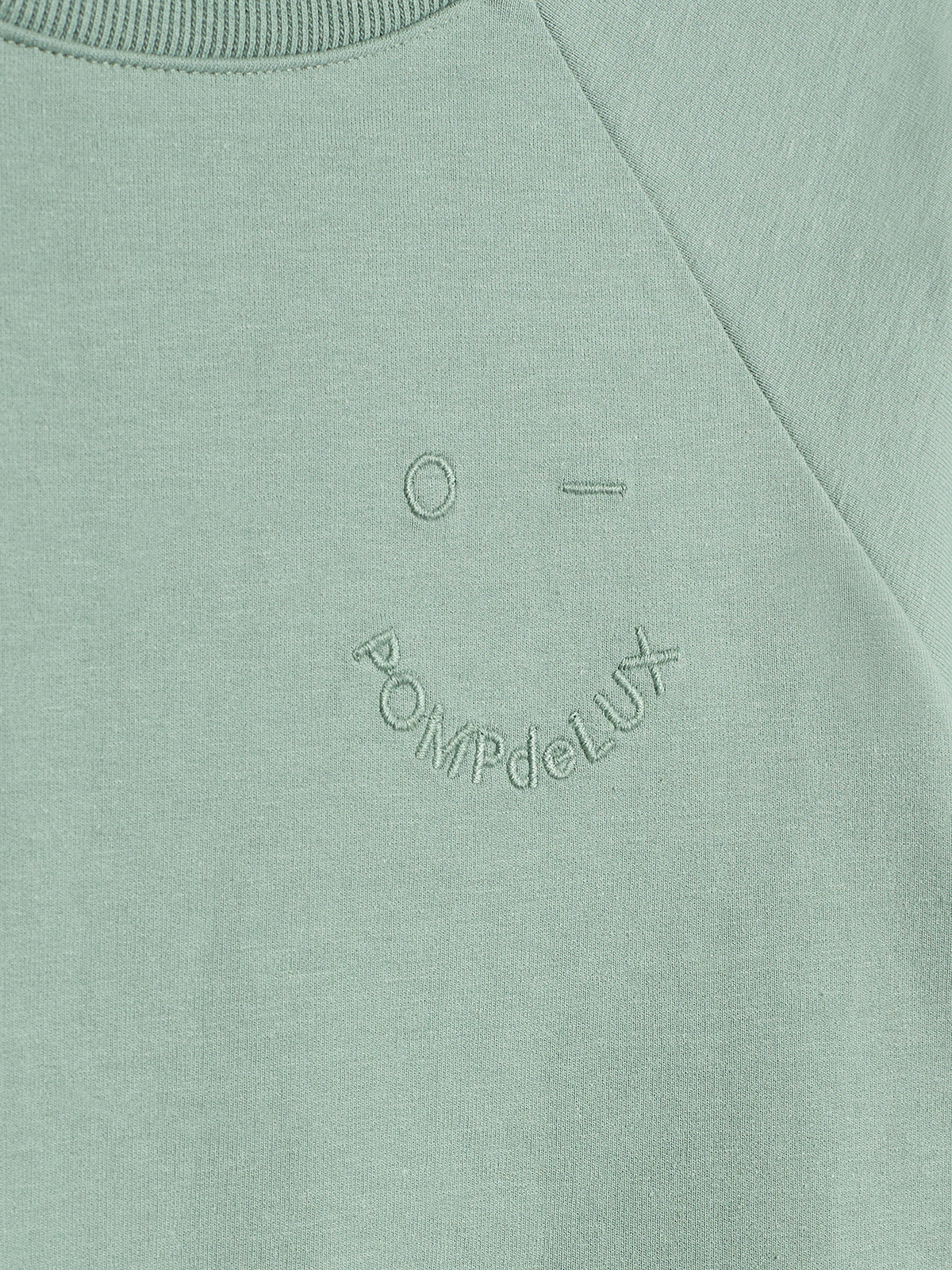 A light green sweatshirt features minimal stitching of a face with "POMPdeLUX" embroidered in a semi-circle below the face. The fabric has a smooth texture with ribbed neckline detail.
