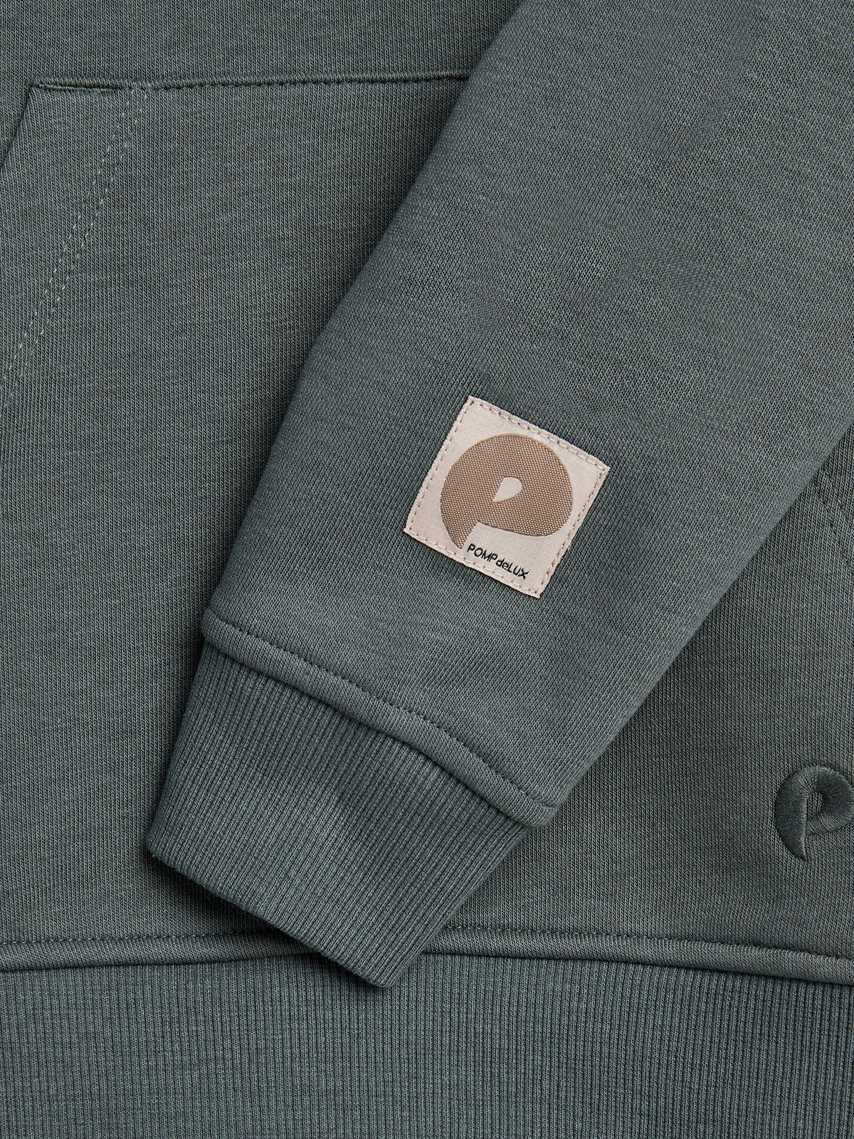 A gray-green sweatshirt displays its ribbed sleeve with a small beige label featuring a stylized "P" and the text "POP delux." It rests against the garment's main body.