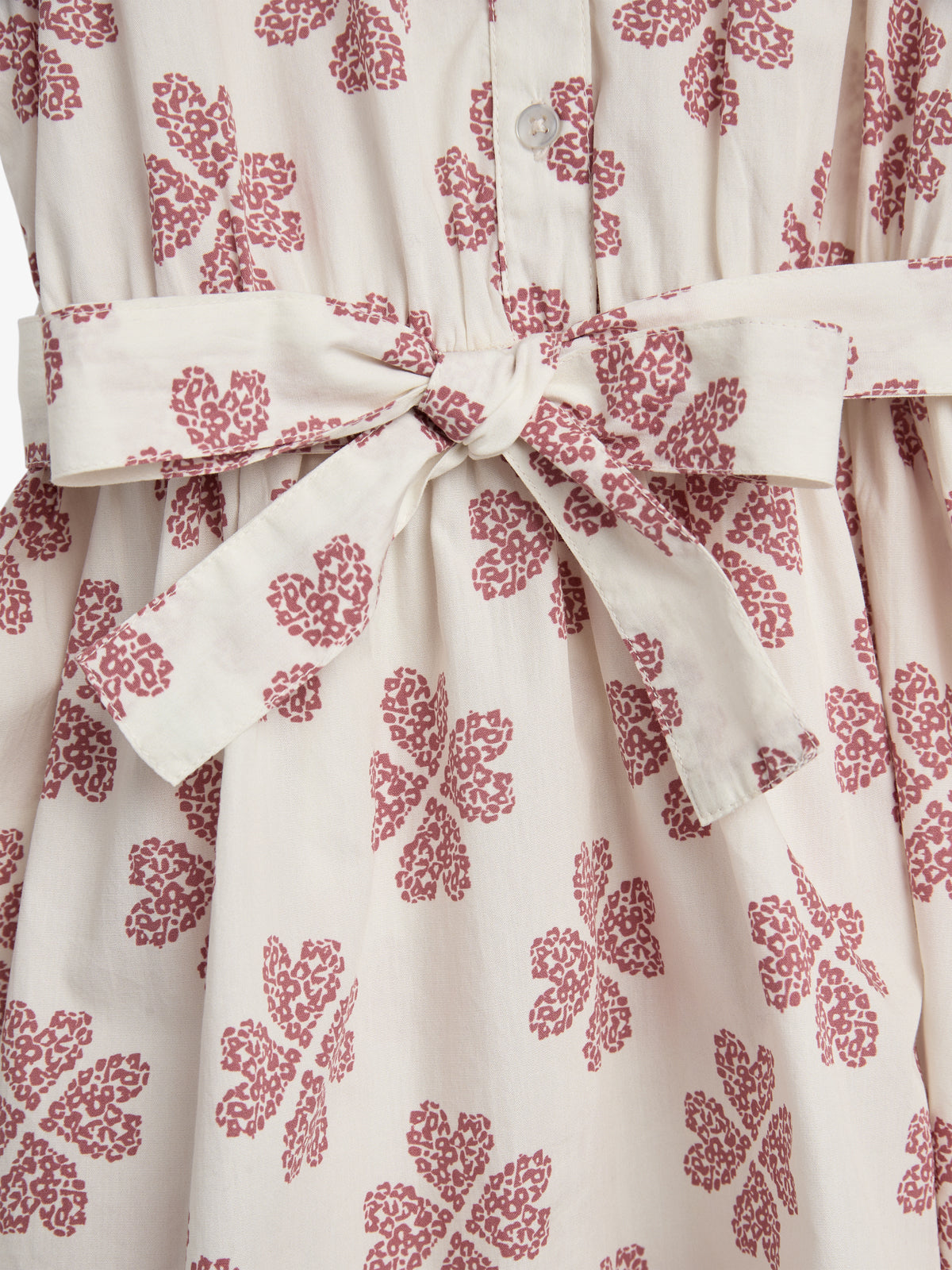 A white dress features a tied bow at the waist and a pattern of reddish floral motifs, with buttons running down the front, displayed against a plain background.