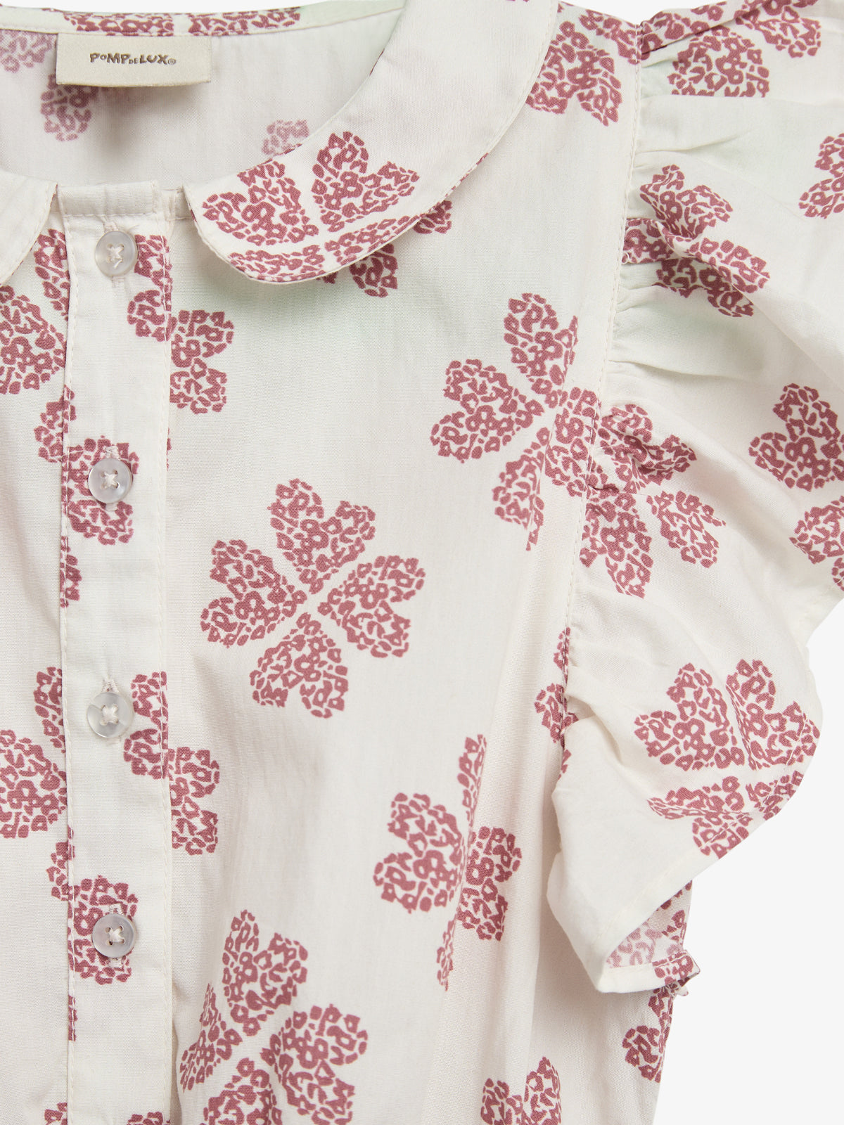 A white blouse with a collar displays pink floral patterns and clear buttons, accompanied by ruffled sleeves. Text reads "POMPdeLUX" on the inside label.