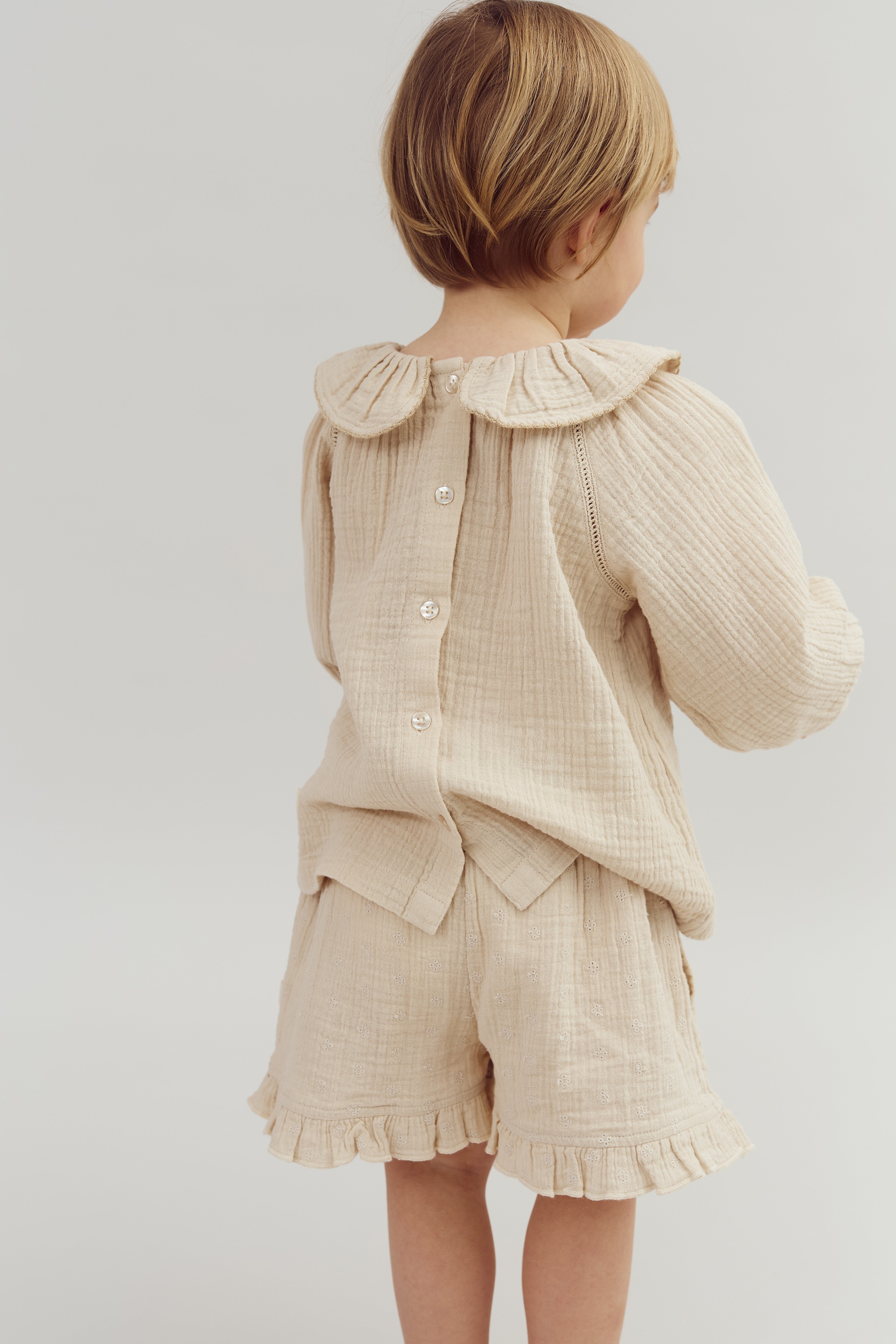 A child stands, wearing a beige, textured outfit with buttoned back and ruffled edges, in a neutral, well-lit setting. The clothing's fabric appears soft and comfortable.