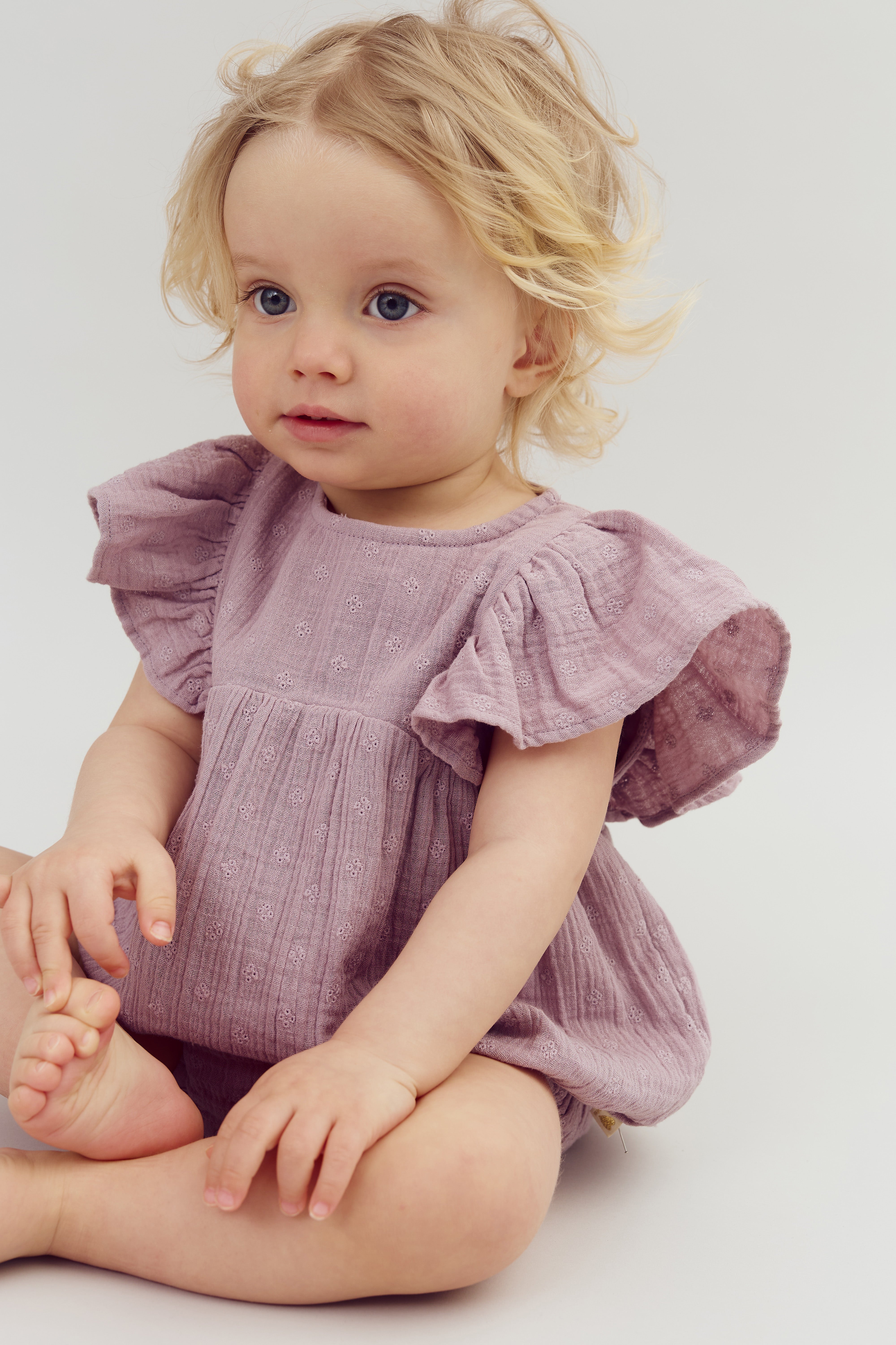 A young child with blonde hair sits cross-legged, wearing a lavender dress with ruffled sleeves. The background is a simple, light-gray studio setting.