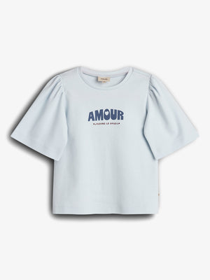 A short-sleeved, light blue shirt with puffed sleeves displays the text "AMOUR" in bold, large letters and "J'ADORE LE SOLEIL" beneath. The shirt is on a plain background.