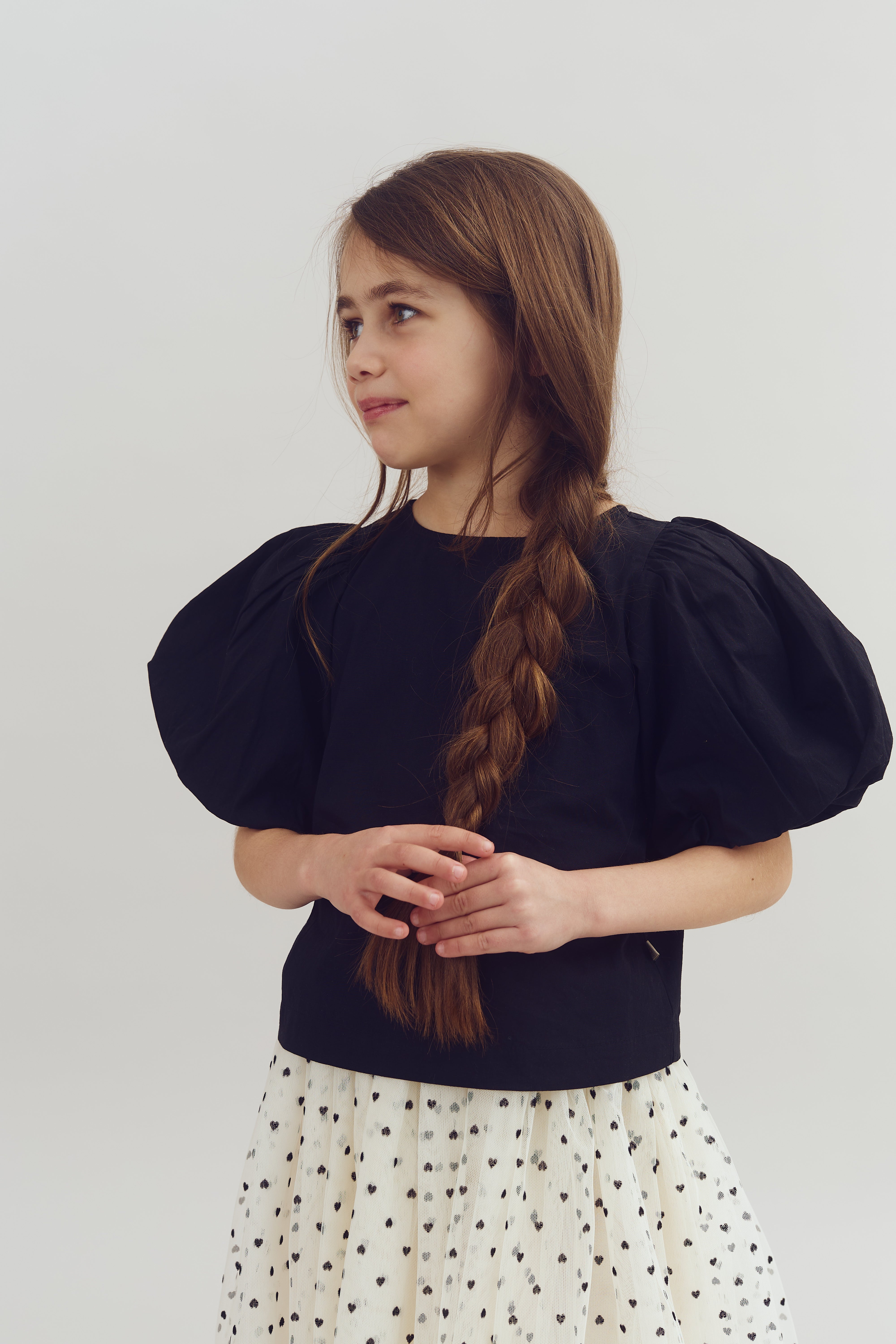 A young girl with long braided hair stands in a neutral setting, wearing a black top with puffed sleeves and a white skirt with black polka dots.