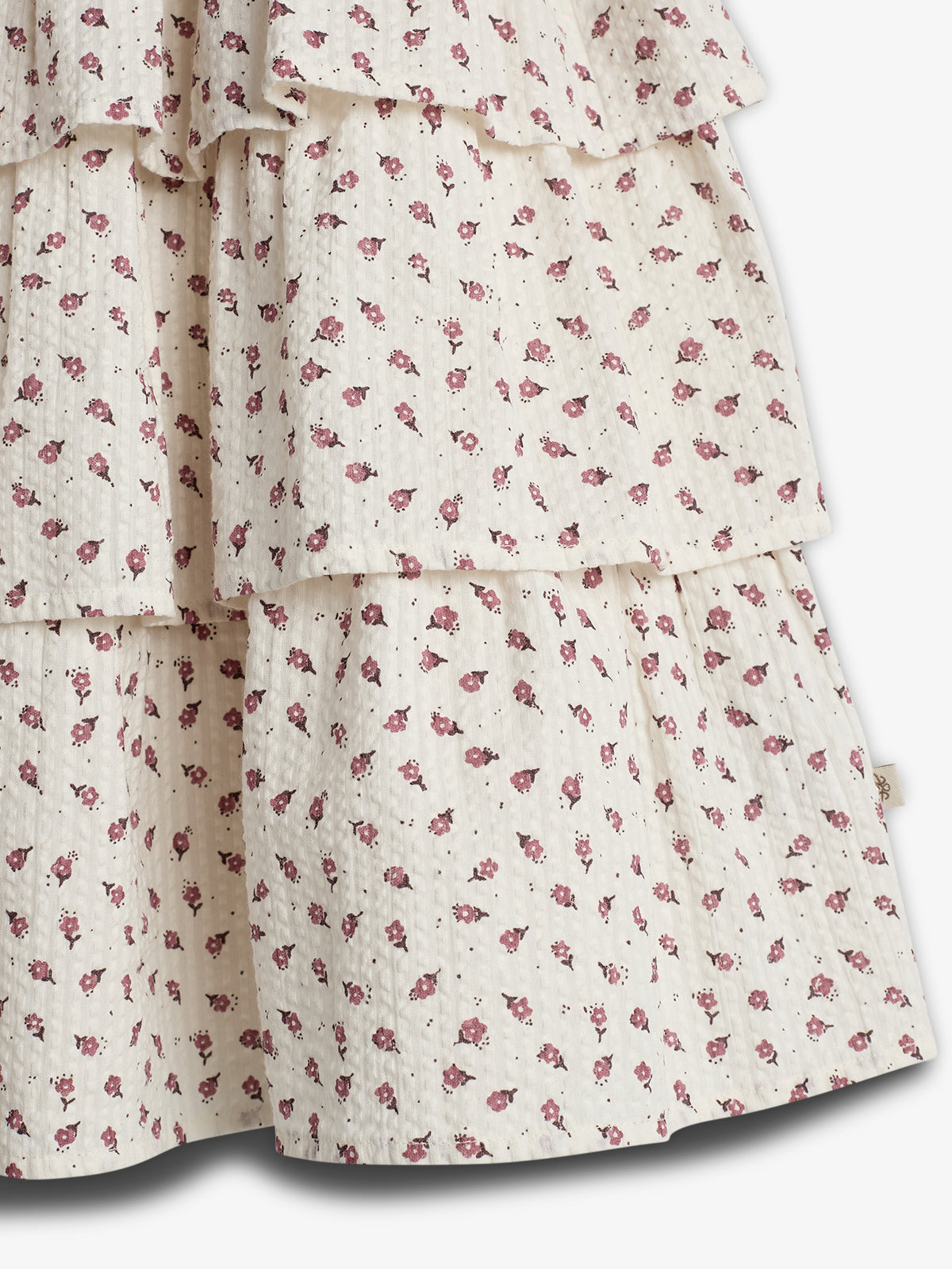 Ruffled fabric layers adorned with small pink floral patterns overlap, creating a textured, tiered design. The background is a light cream color, adding a soft, delicate appearance.