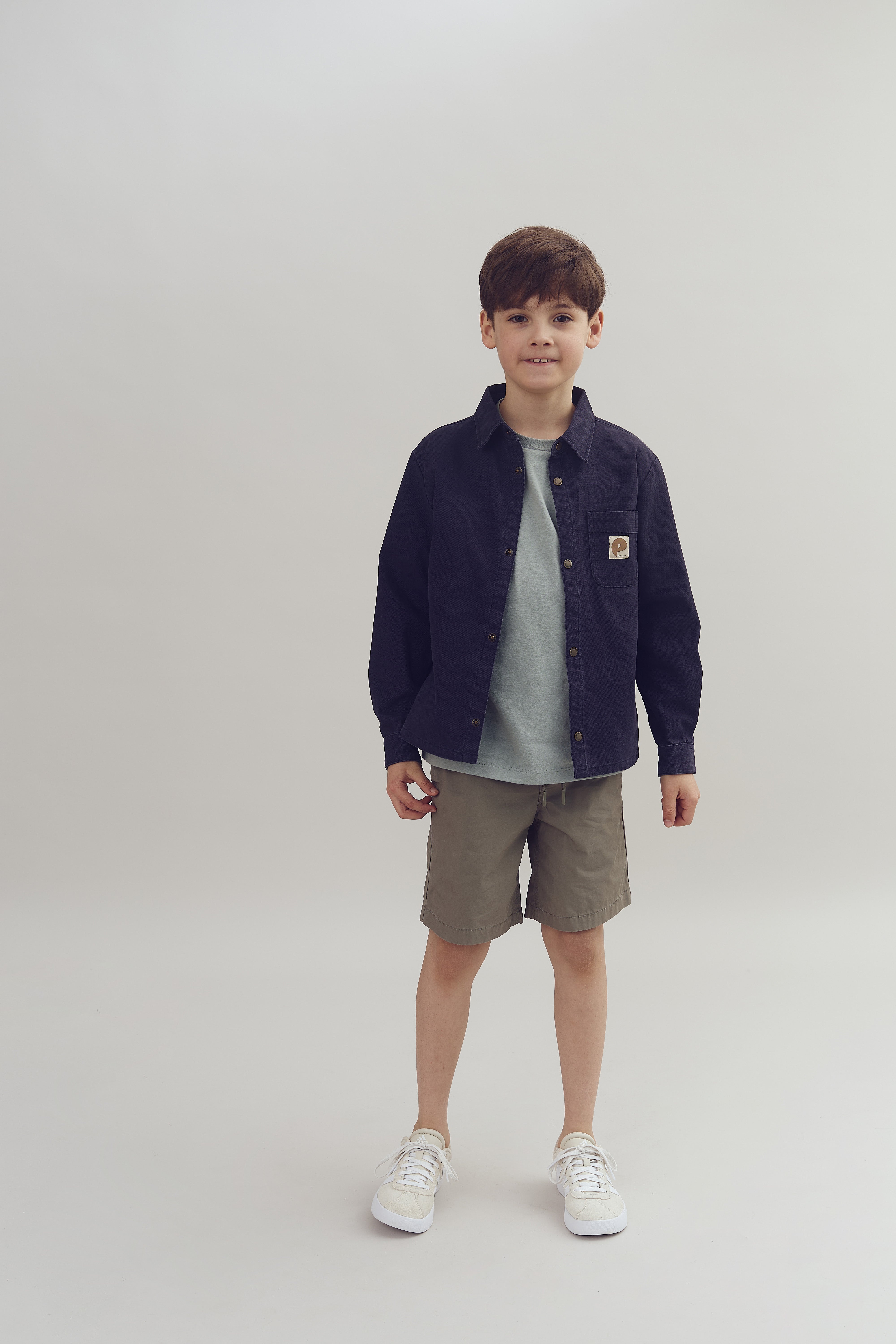 A young boy smiles, standing confidently in a minimalist studio. He wears a navy jacket over a gray shirt, khaki shorts, and white sneakers, with a beige logo patch on the pocket.