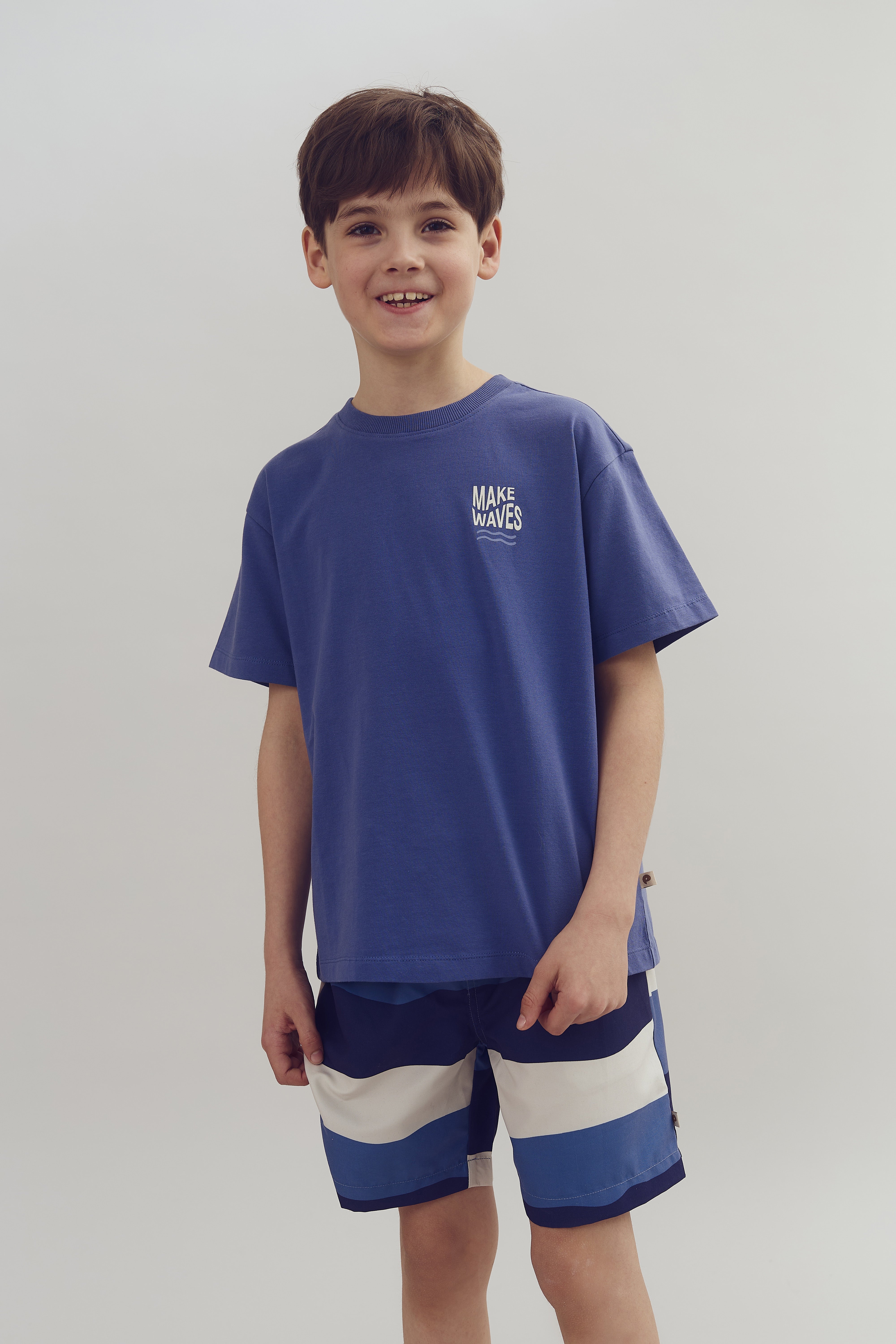 A child stands smiling, wearing a blue T-shirt with "MAKE WAVES" text and striped shorts. The background is plain white, emphasizing the clothing.