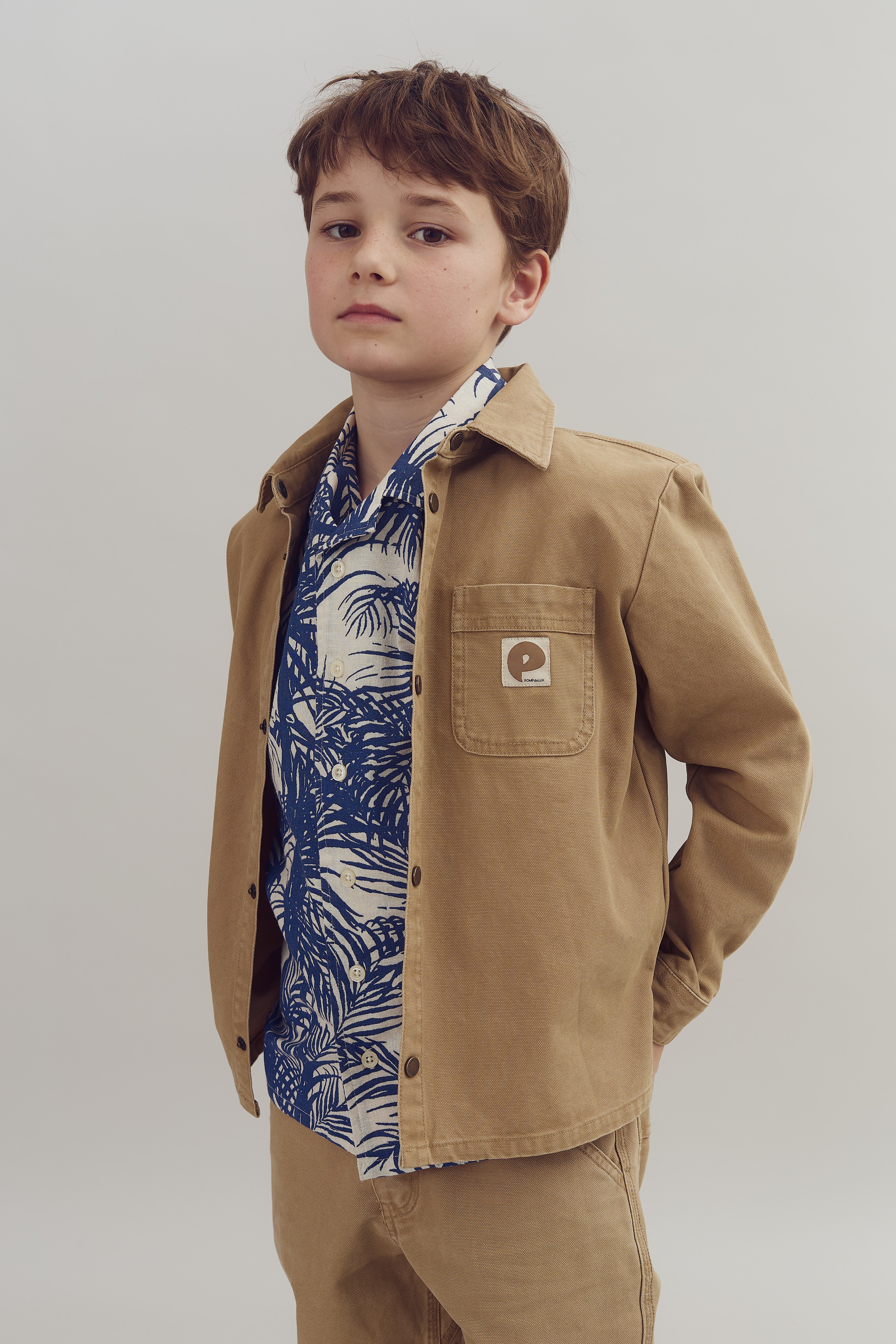 A child wearing a tan jacket with the letter "P" on the chest pocket stands confidently, hands in pockets. The blue and white patterned shirt adds a stylish touch against a neutral background.