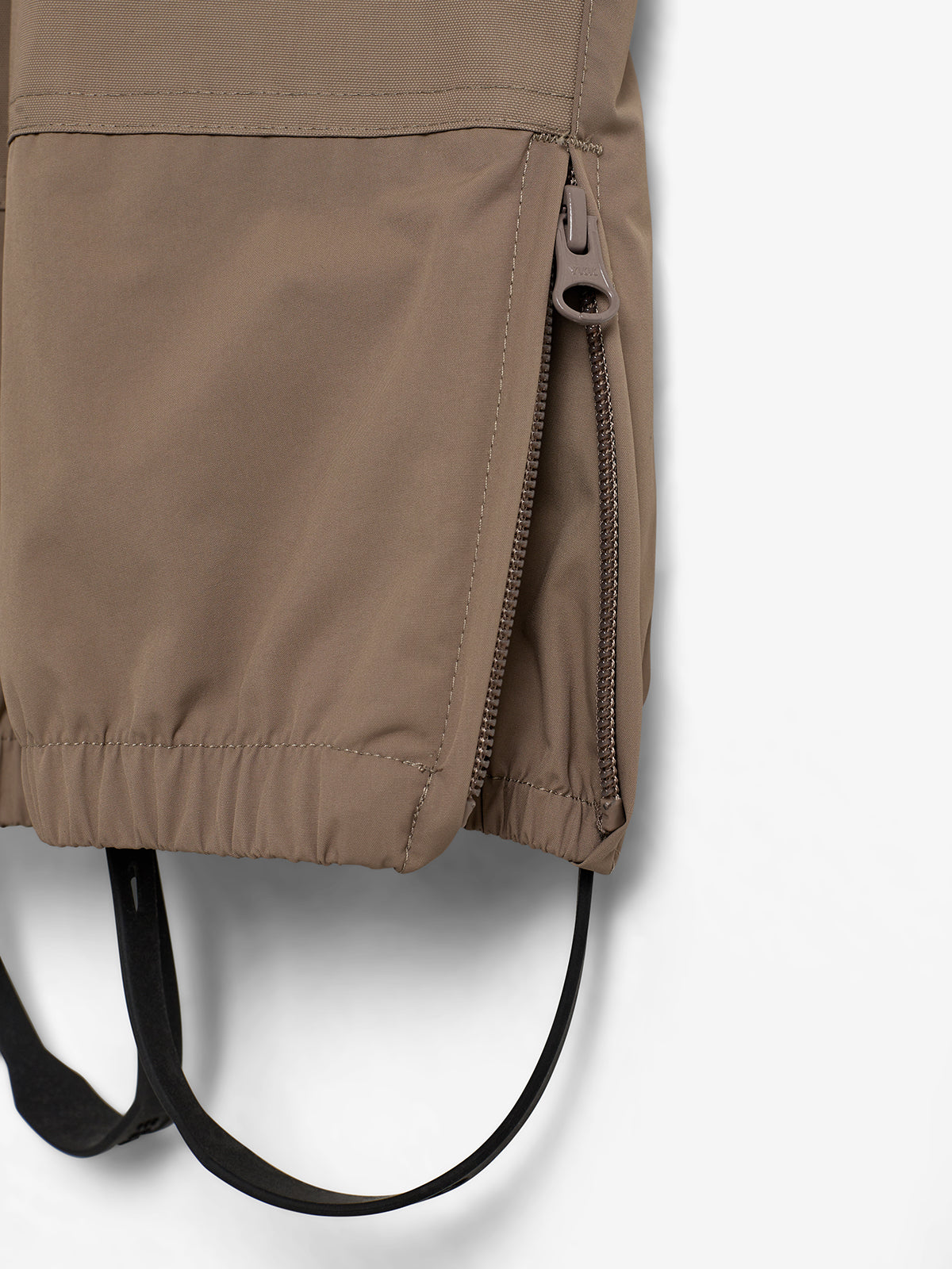 Braune Jacke mit einem Reißverschluss an der Seite, der leicht geöffnet ist. Elastische Schlaufe hängt darunter, in neutralem Hintergrund.