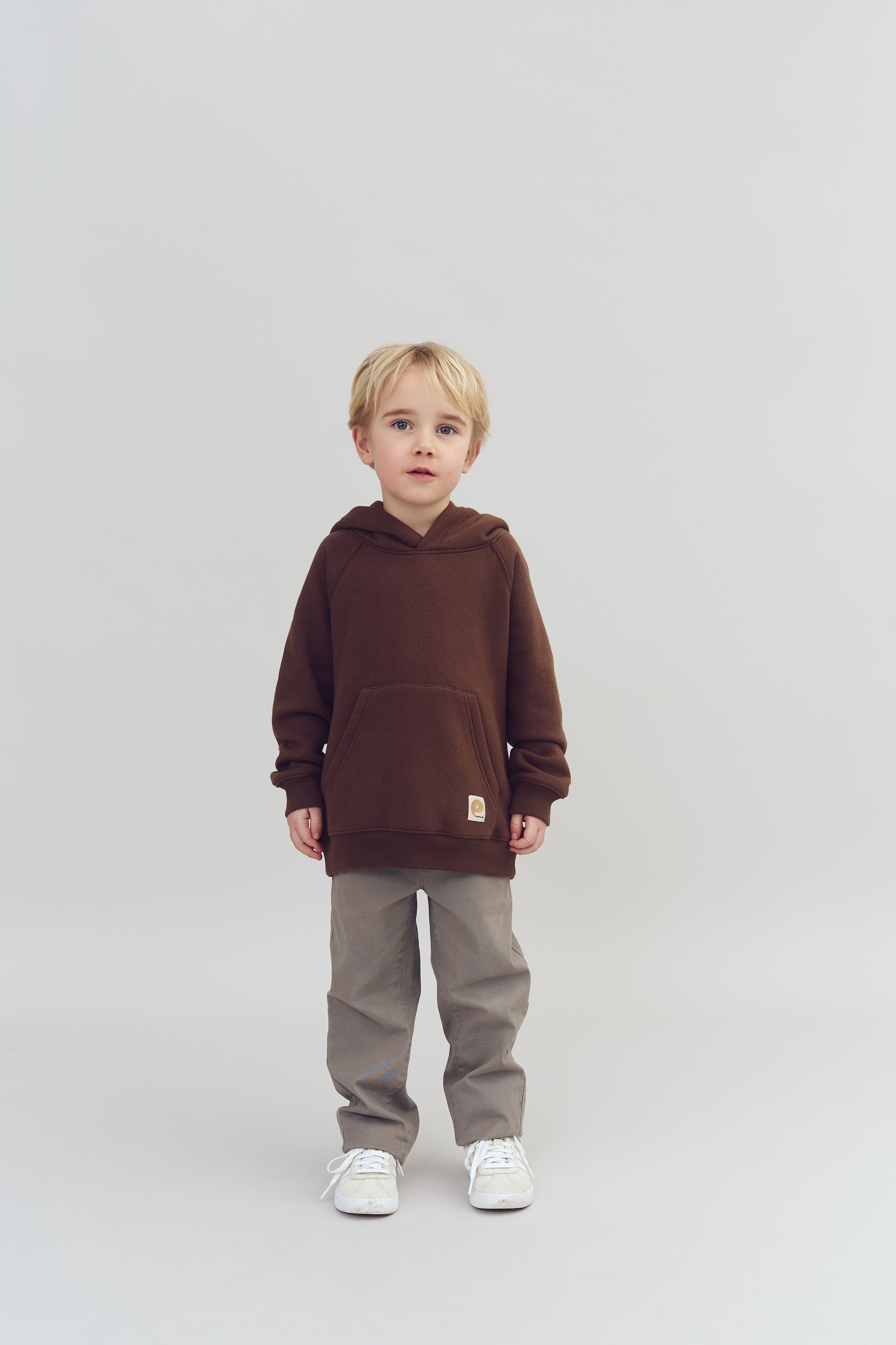 Ein kleines Kind, das einen braunen Hoodie und graue Hosen trägt, steht still auf einem hellen Hintergrund. Es schaut direkt in die Kamera.