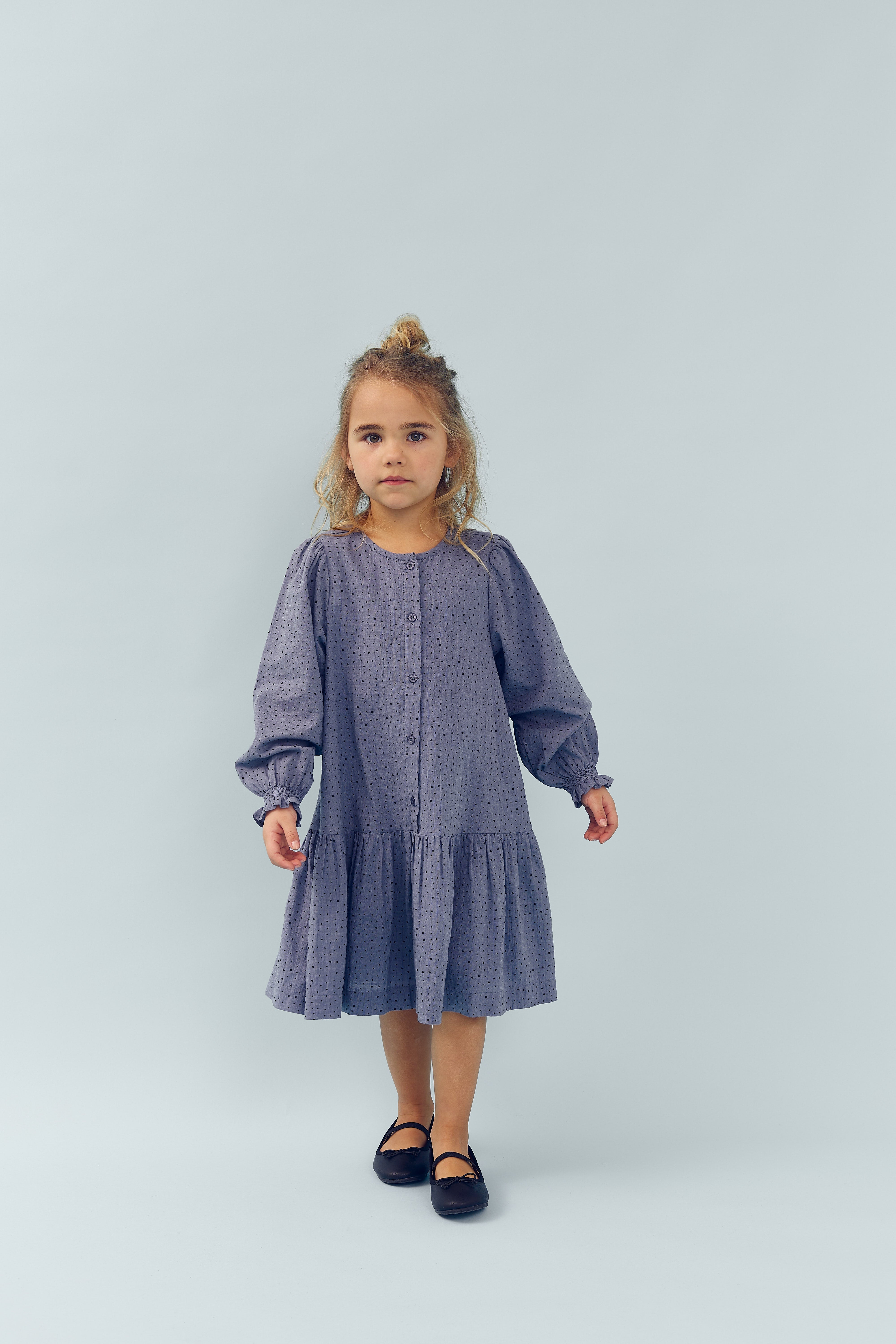 A young child stands in a light blue setting, wearing a long-sleeved, patterned purple dress and black shoes, looking directly ahead.