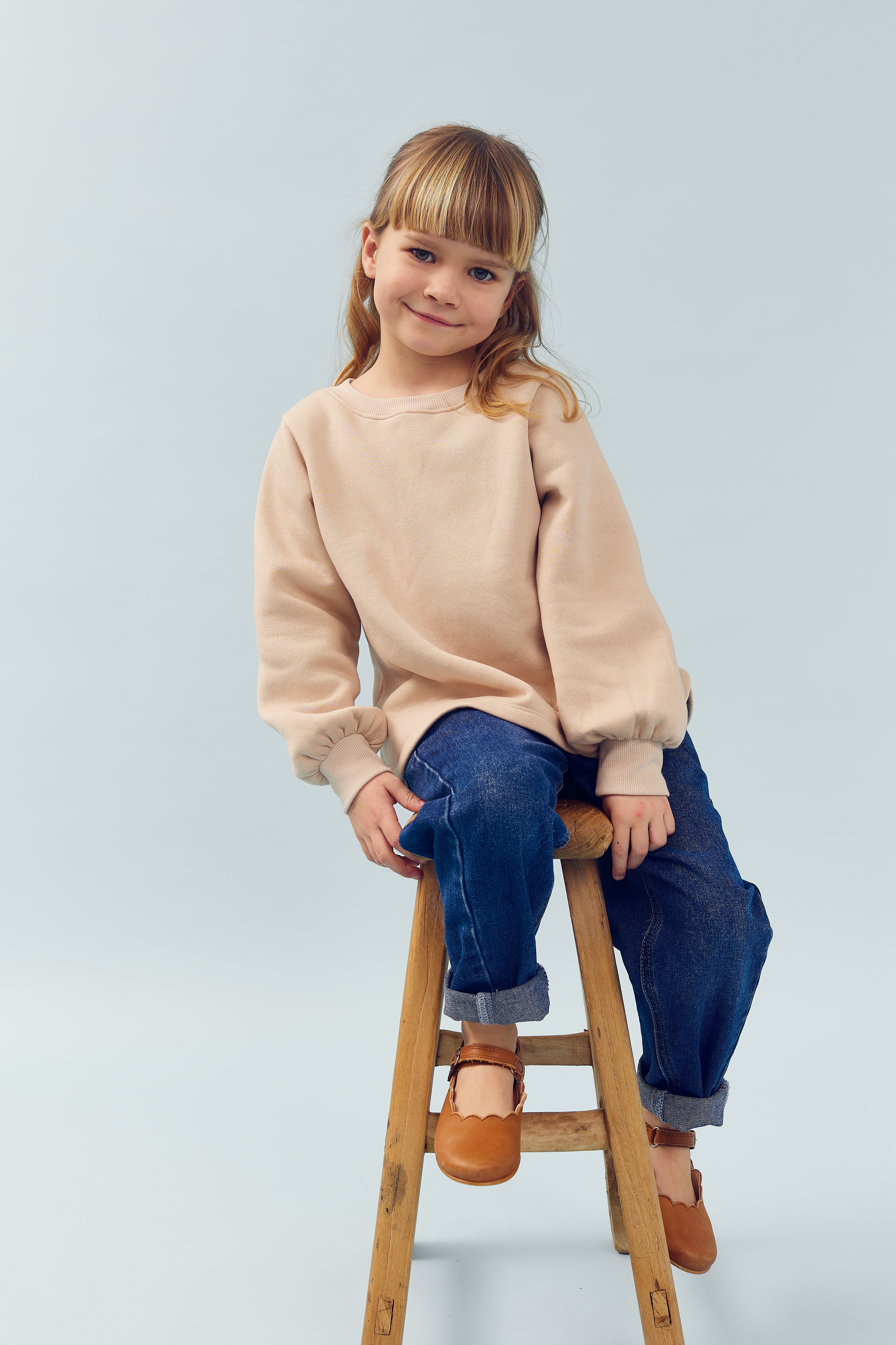 Ein Kind sitzt lächelnd auf einem Holzhocker. Es trägt einen beigefarbenen Pullover, blaue Jeans und braune Schuhe. Der Hintergrund ist einfarbig hellblau.