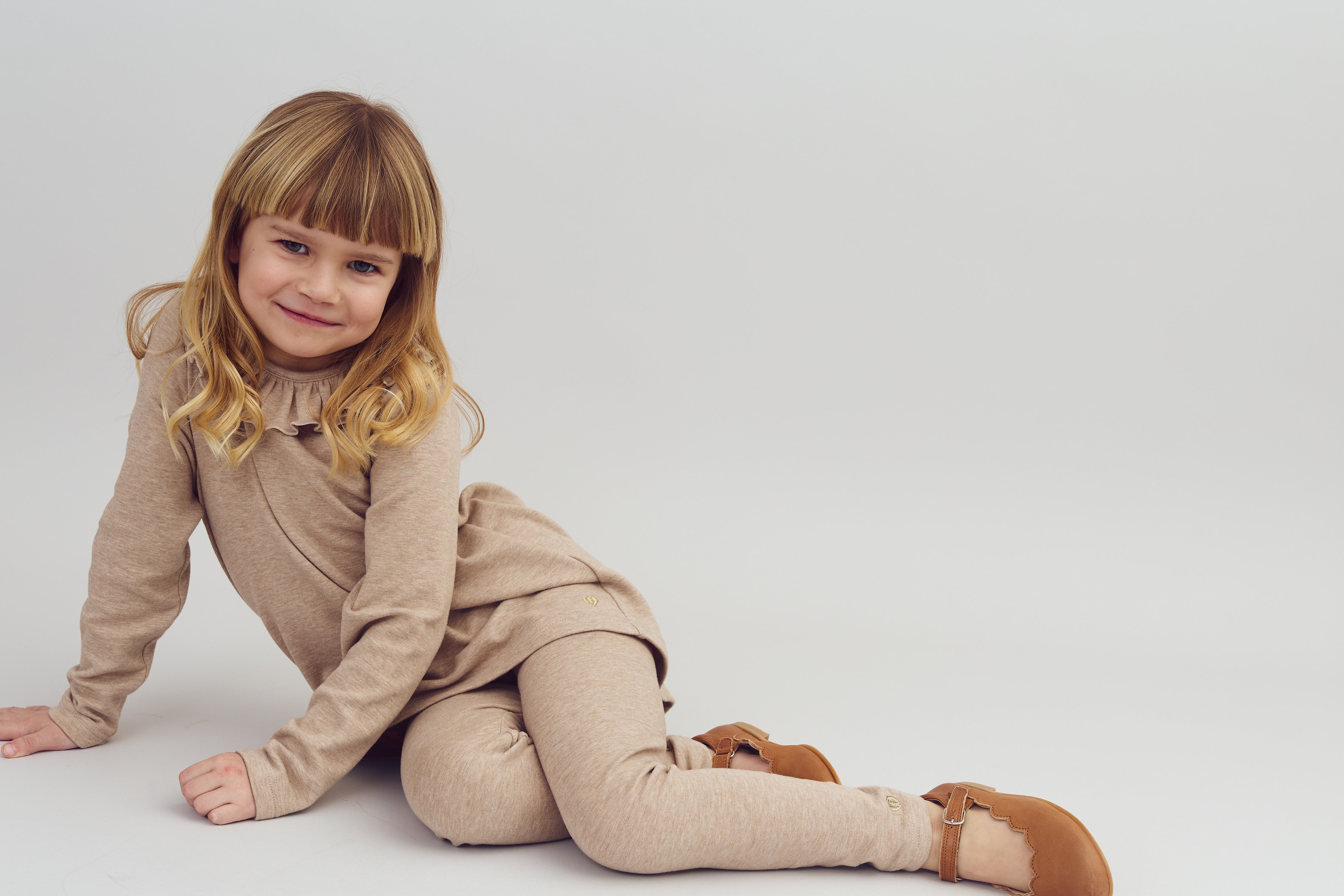 A young child, wearing a beige outfit and brown shoes, sits on the floor with one hand resting on the ground, smiling against a plain gray background.