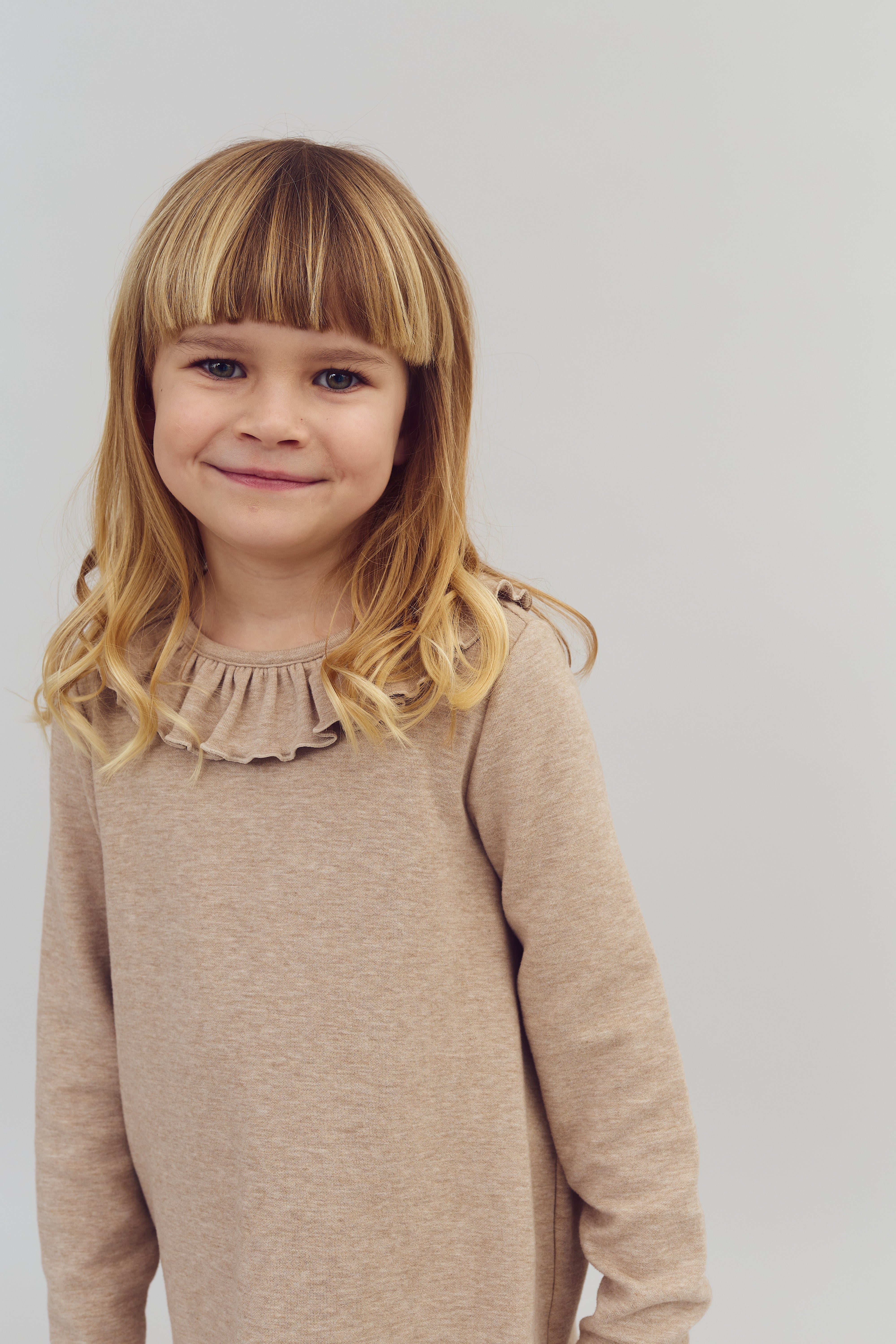 A young child with blond hair is smiling gently, wearing a beige sweater with a frilled collar; the background is plain and light gray.