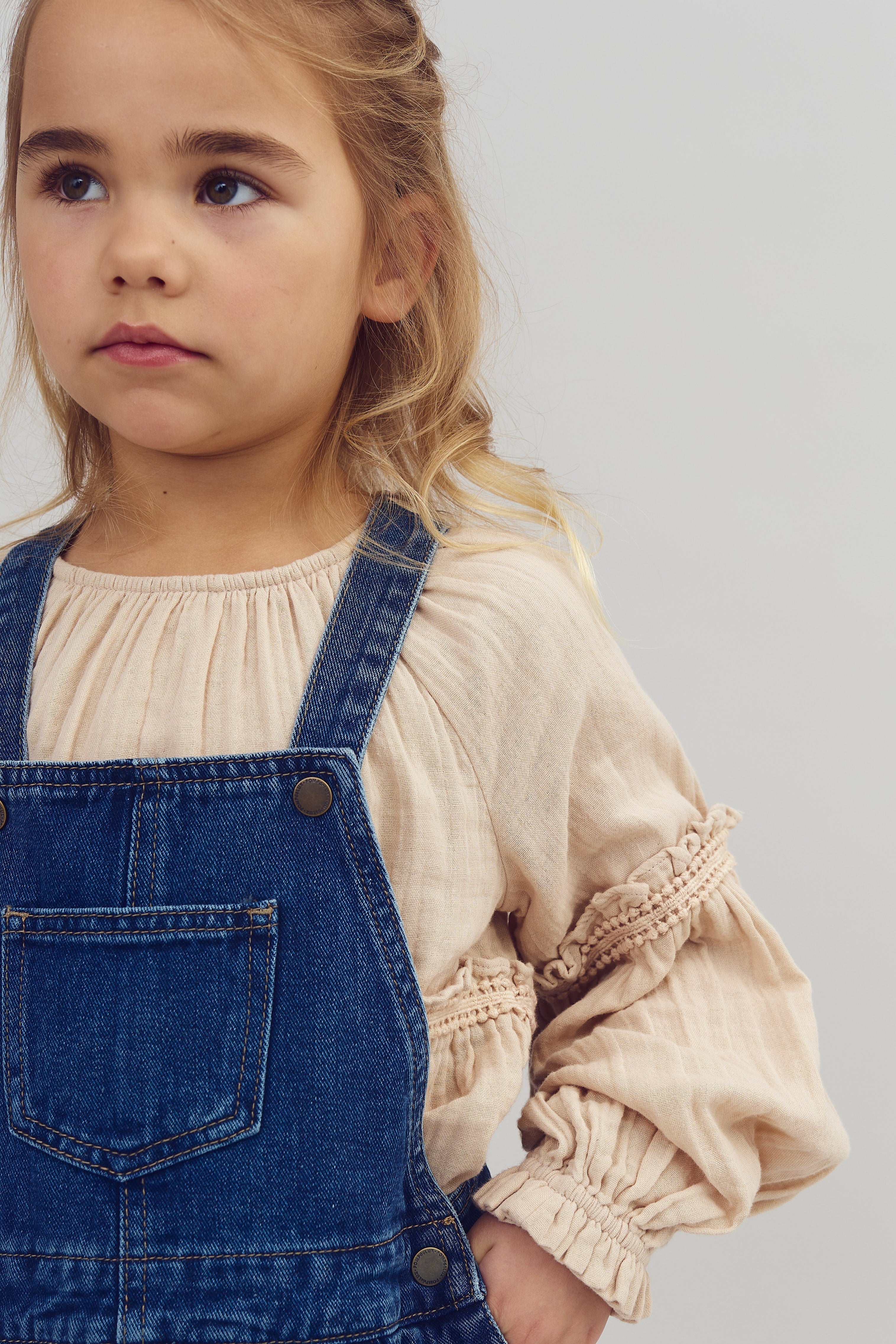 Ein Kind mit blondem Haar trägt eine beigefarbene Bluse und eine blaue Latzhose, schaut leicht zur Seite vor einem neutralen, hellen Hintergrund.