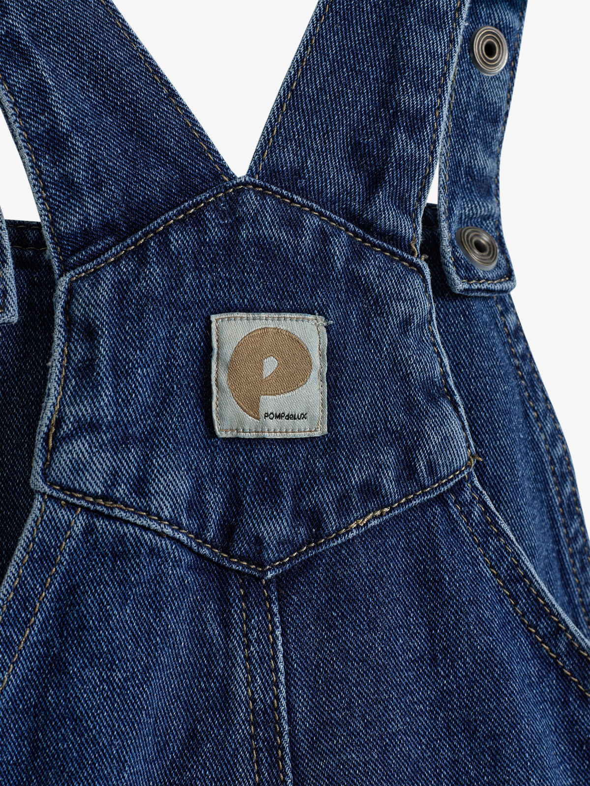 Denim-Träger mit Knopfdetail, sitzen an einer Jeanslatzhose. Ein Quadrat-Etikett zeigt ein "P" und den Text "POMPdeLUX". Steppnähte umranden die Kanten. Umgebung nicht sichtbar.
