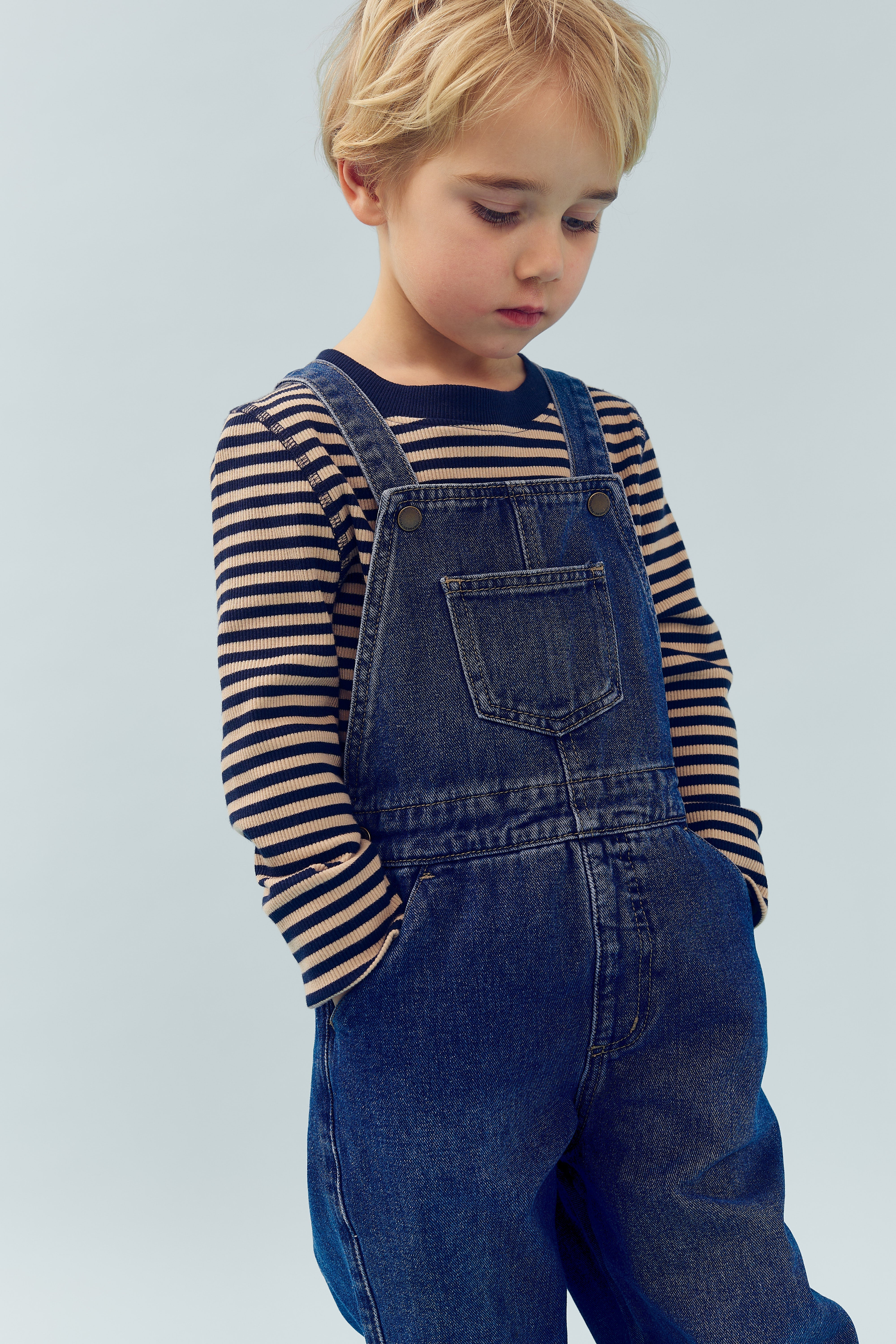 Kind zeigt traurigen Ausdruck, trägt gestreiftes Langarmshirt und blaue Jeanslatzhose. Hände in den Taschen, neutraler graublauer Hintergrund.