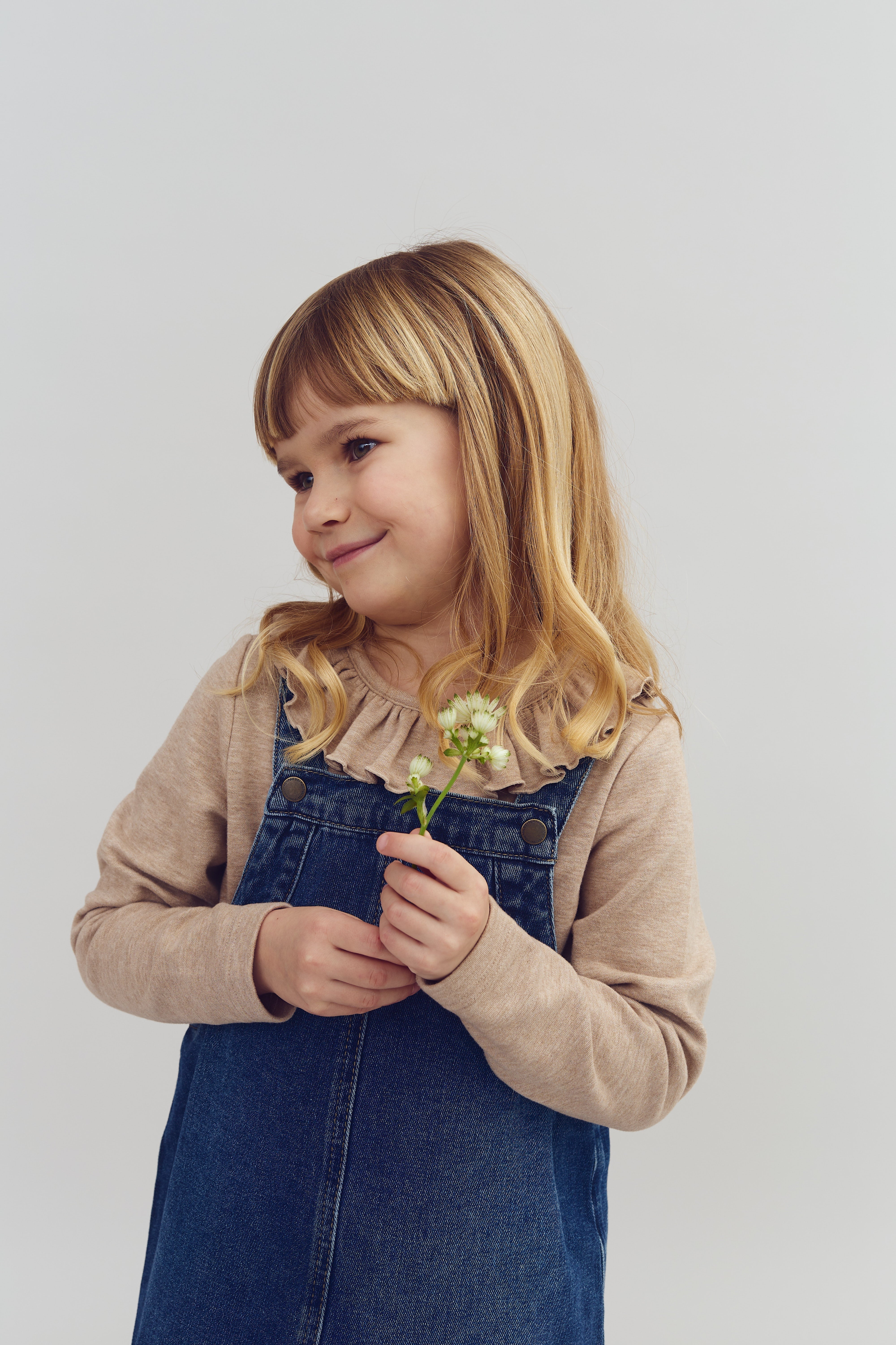 A young child, holding a small white flower, smiles softly while wearing a denim dress and beige top against a plain light gray background.