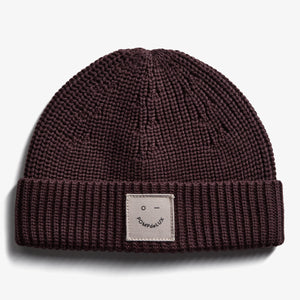A knitted maroon beanie with a folded rim features a beige patch labeled "POMPdeLUX" and a simple face design, resting on a plain white background.