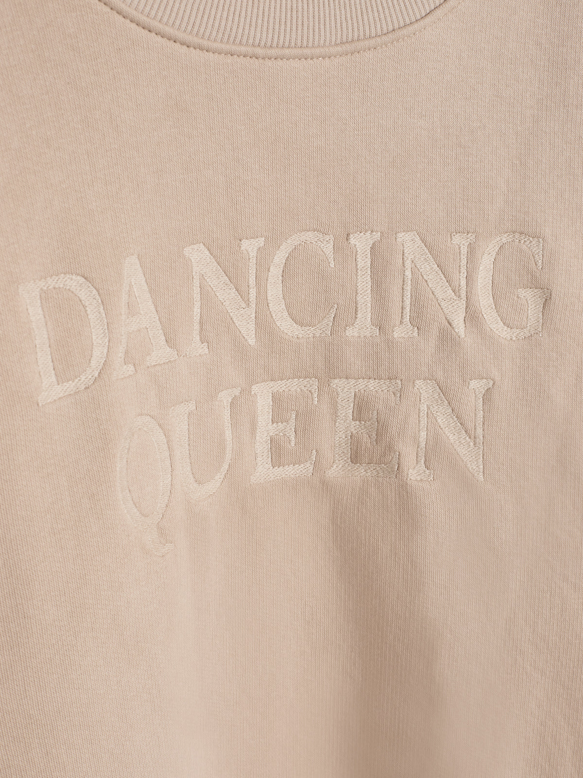 Ein beigefarbener Pullover zeigt die Textstickerei "DANCING QUEEN". Der Stoff hat einen gerippten Kragen und das Gesamtbild wirkt gemütlich und stilvoll.