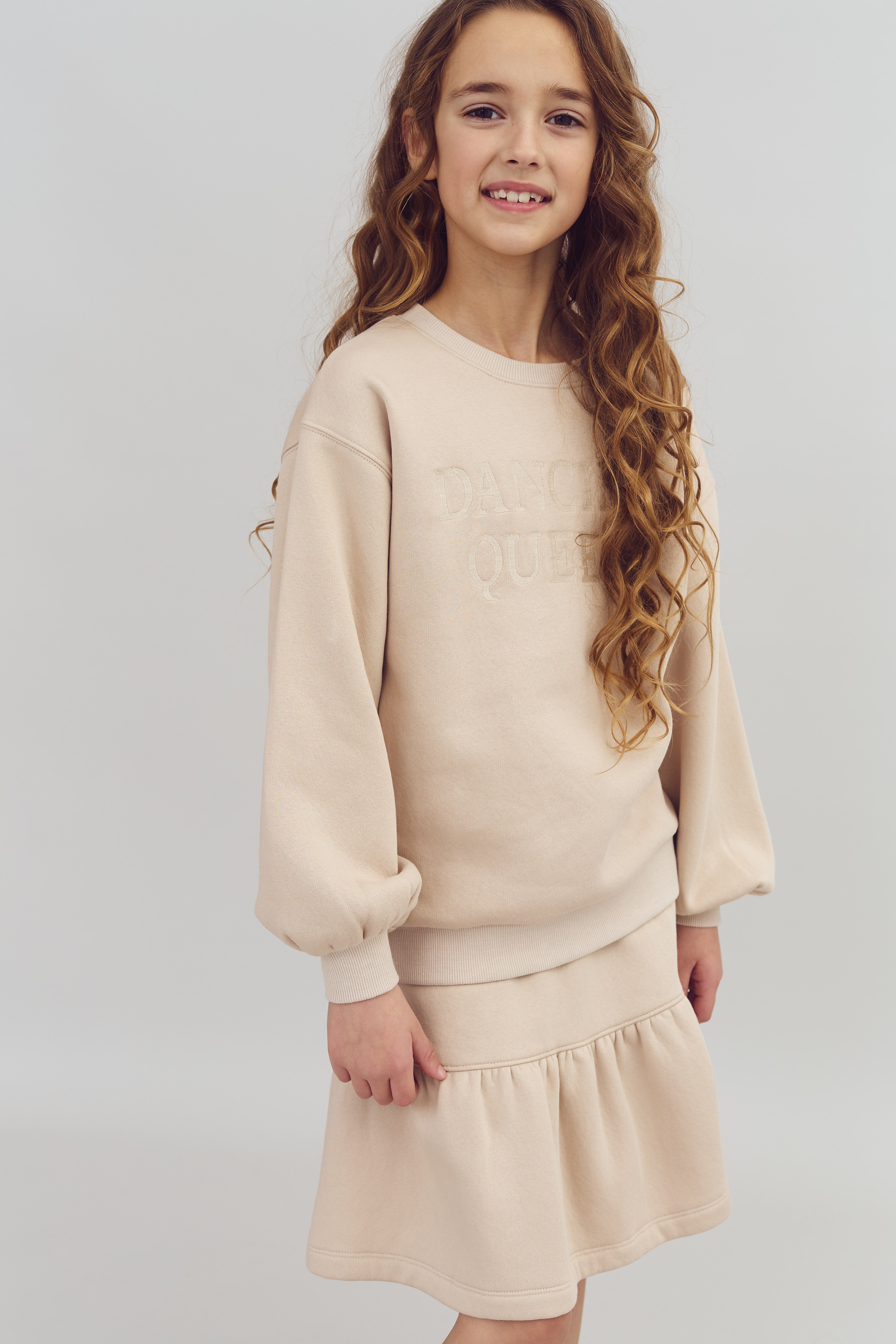 Mädchen lächelt, trägt einen beigen Pullover mit dem Text "DANCE QUEEN" und einen passenden Rock, stehend vor neutralem Hintergrund.