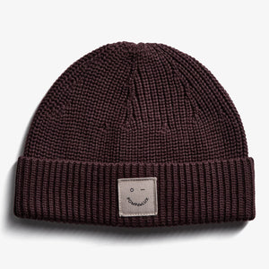 A knitted beanie resting flat on a white surface features a visible patch with a smiley face and "POMPdeLUX" text on the folded brim.