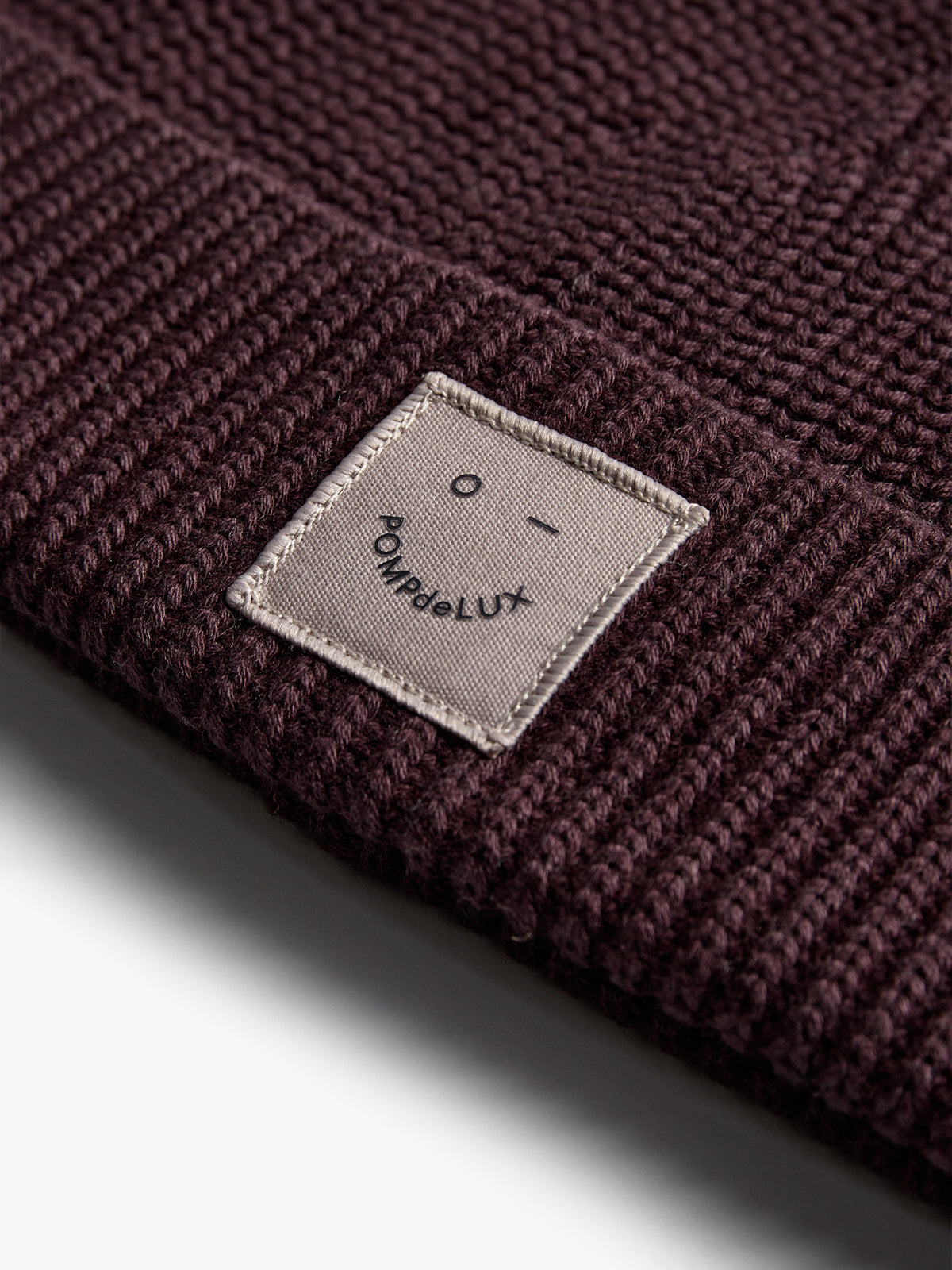 A dark knit beanie displays a sewn label with a minimalist smiley face and the text "POMPdeLUX" against a light background, resting on a flat surface.
