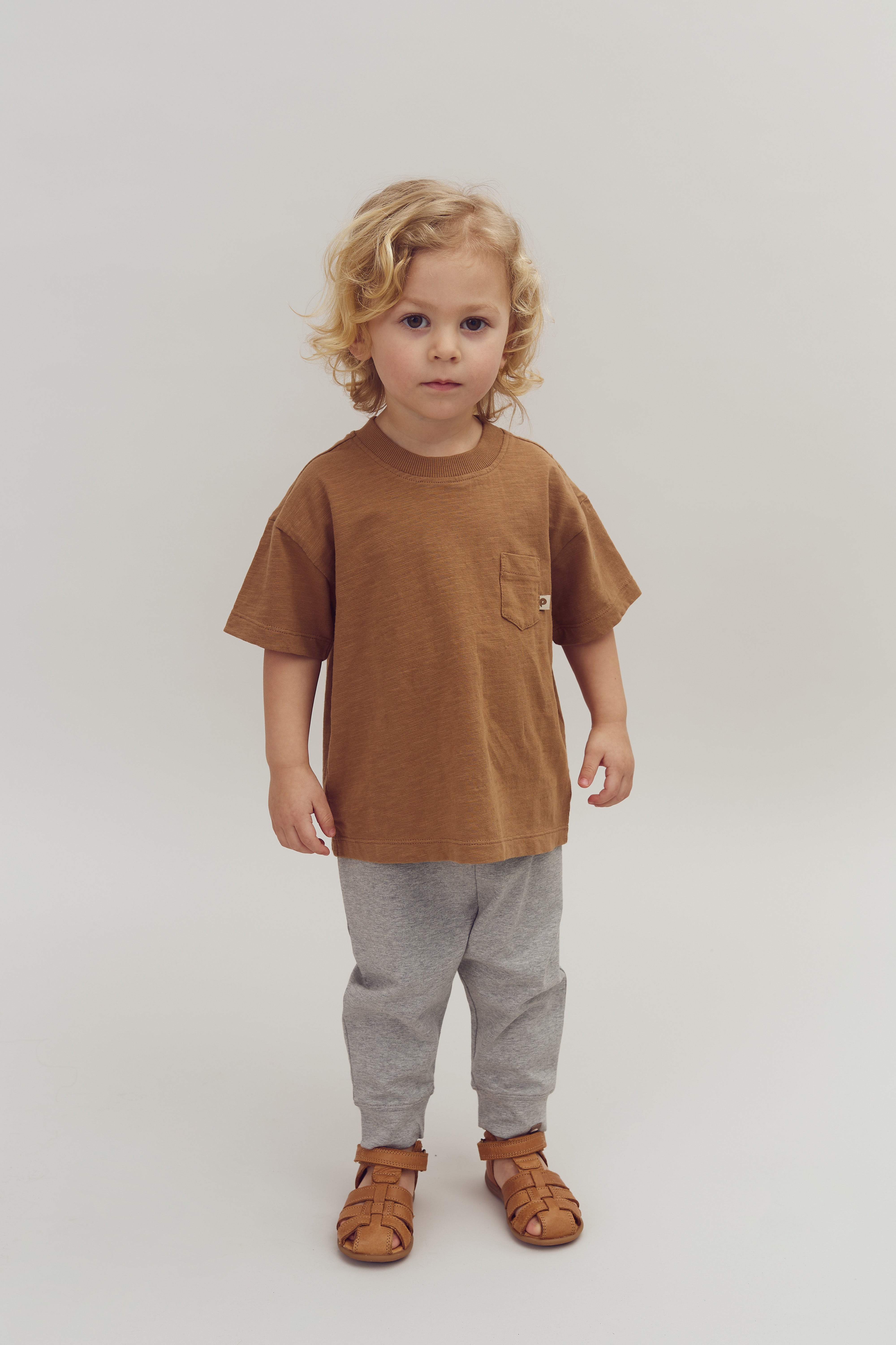 Ein Kind mit blonden Locken steht still. Es trägt ein braunes T-Shirt, graue Hosen und braune Sandalen. Der Hintergrund ist schlicht und weiß.
