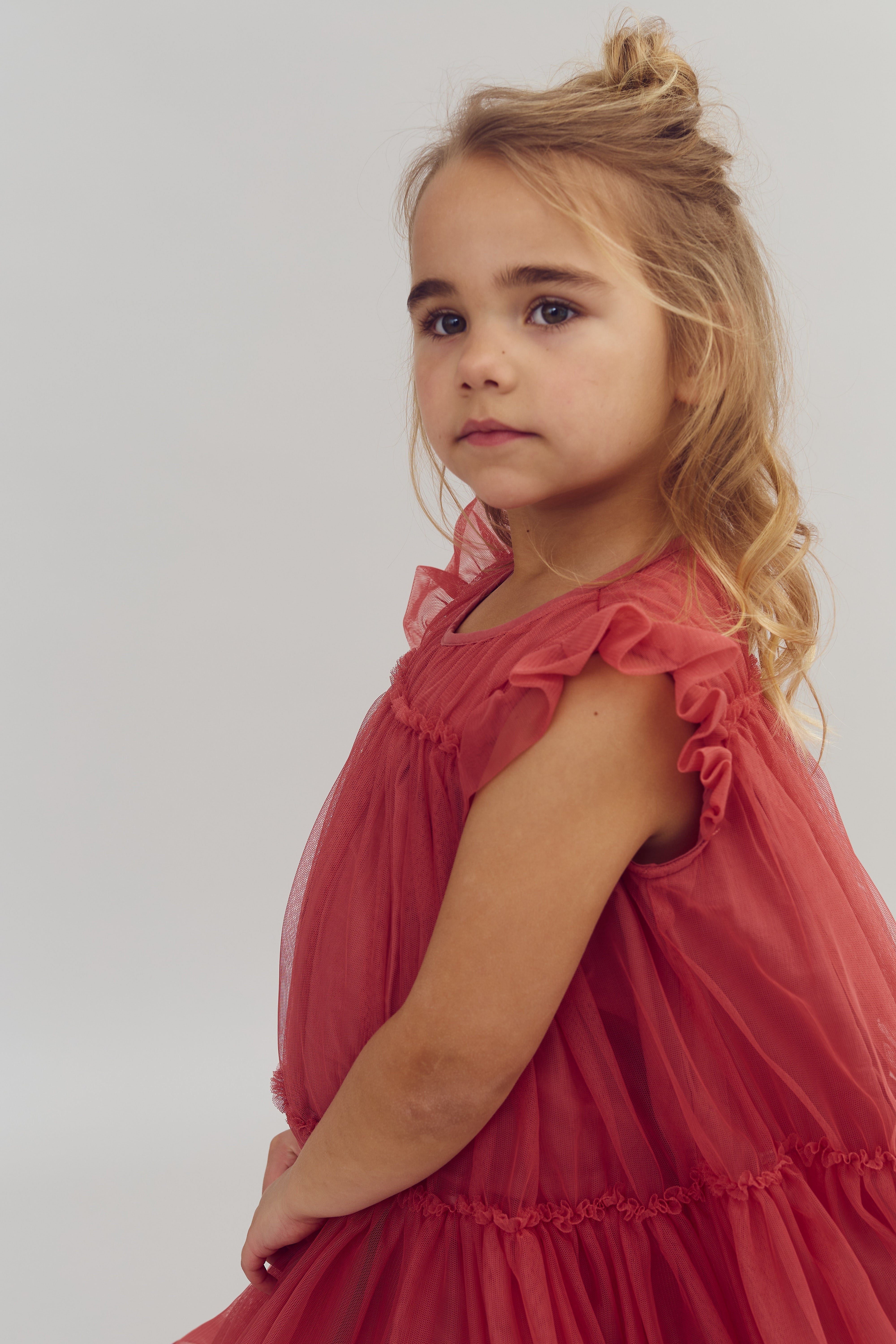 A young girl stands looking into the distance, wearing a red dress with ruffled sleeves. Her light hair is styled in waves and a small topknot, set against a neutral background.