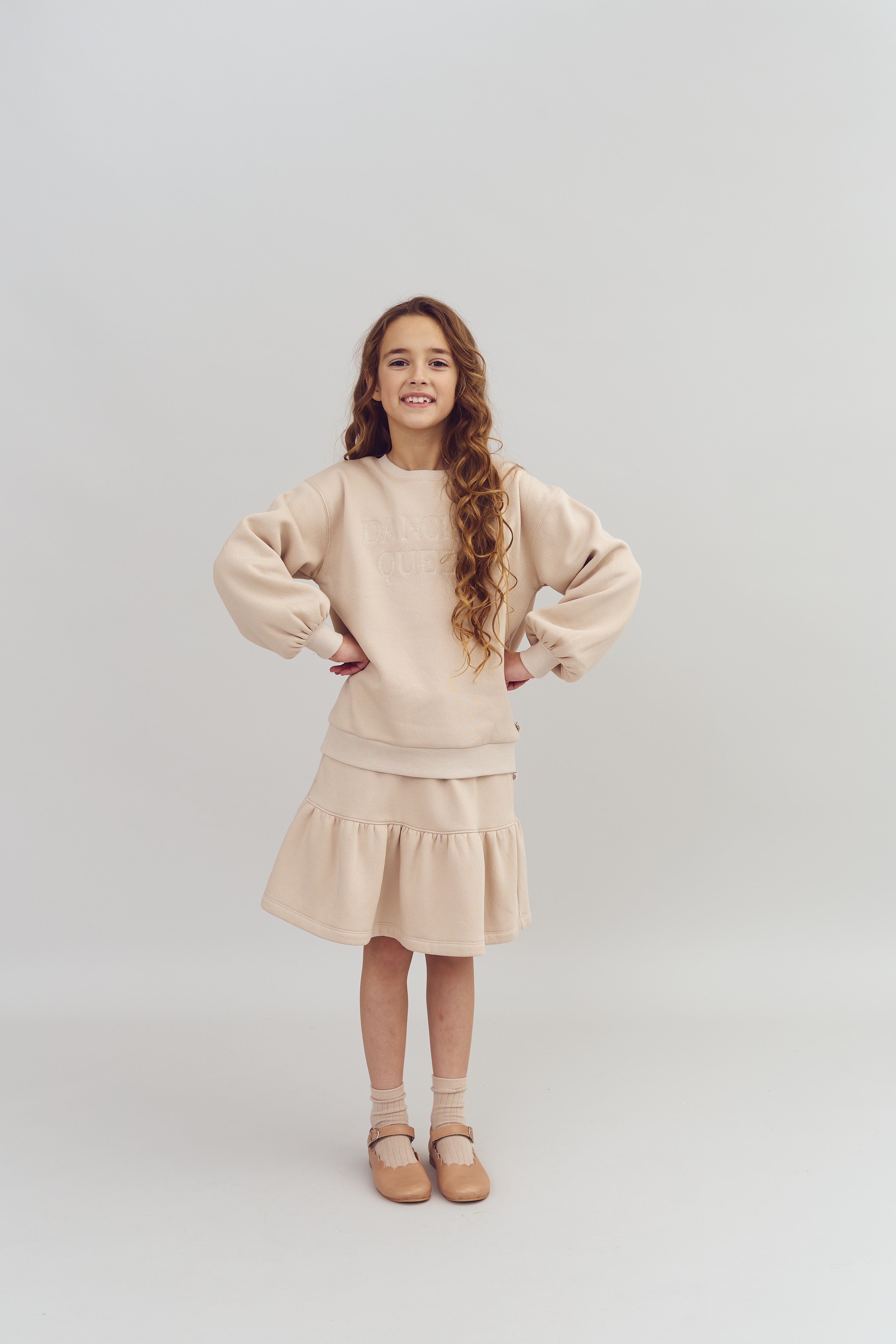 Ein Mädchen im beigefarbenen Outfit steht lächelnd mit Händen in die Hüften gestemmt vor einem neutralen Hintergrund. Auf ihrem Pullover steht "Dancer Queen".