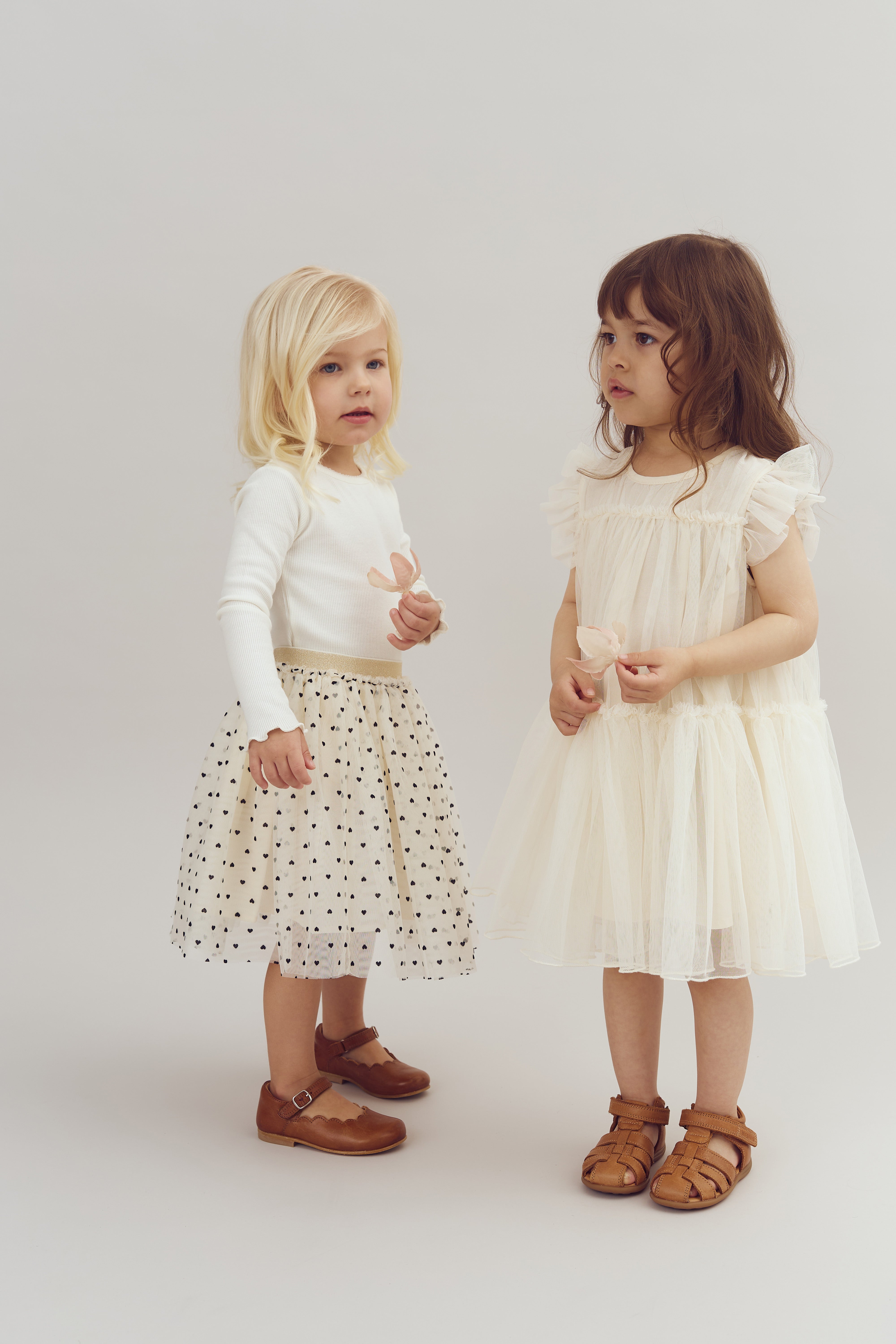 Two young children stand holding flowers. The left child wears a white sweater and polka dot skirt, while the right child wears a frilly, cream dress. Both are in a neutral studio setting.