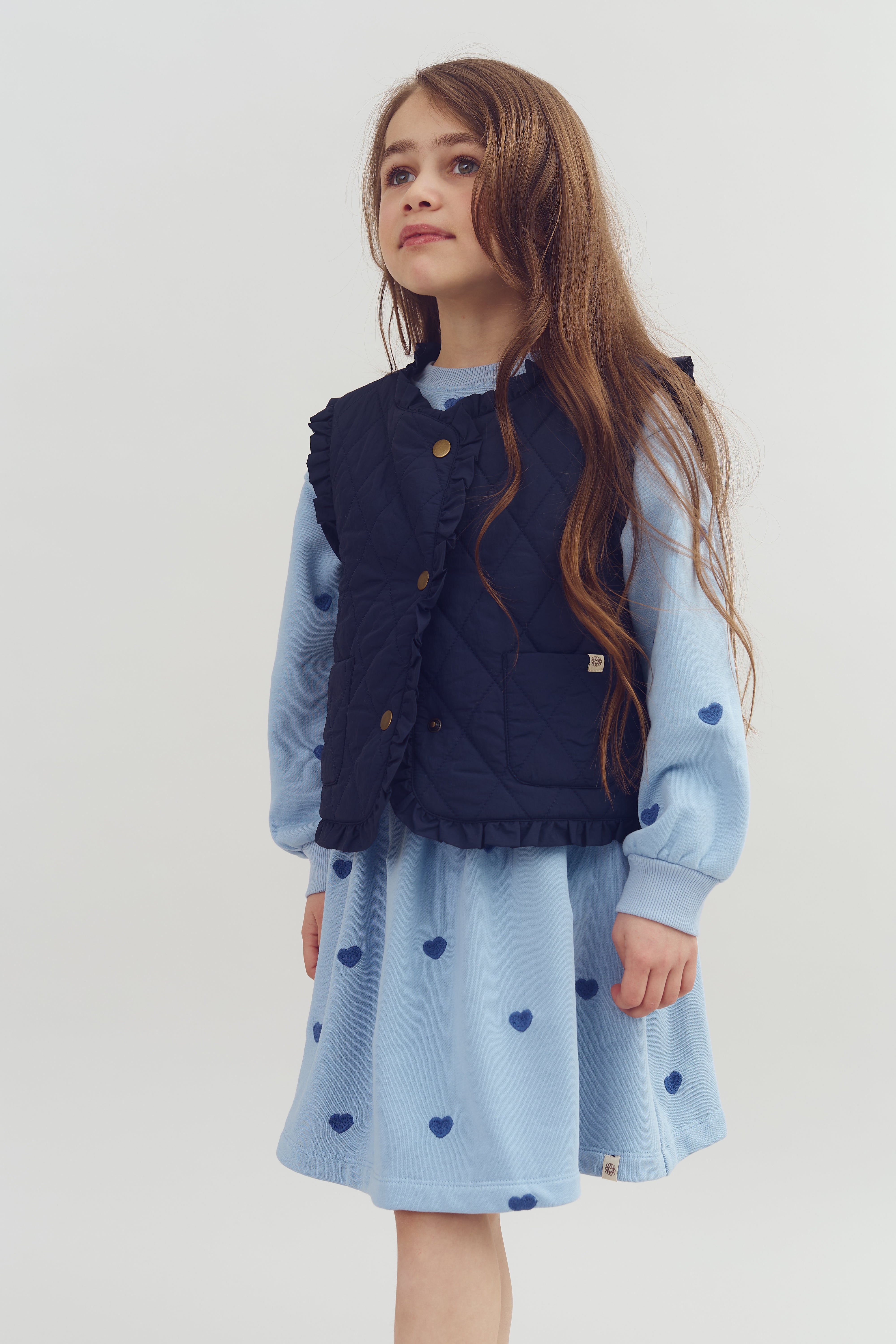 A young girl wearing a quilted navy vest over a light blue dress with blue heart patterns stands gazing upward. She has long brown hair against a plain white background.