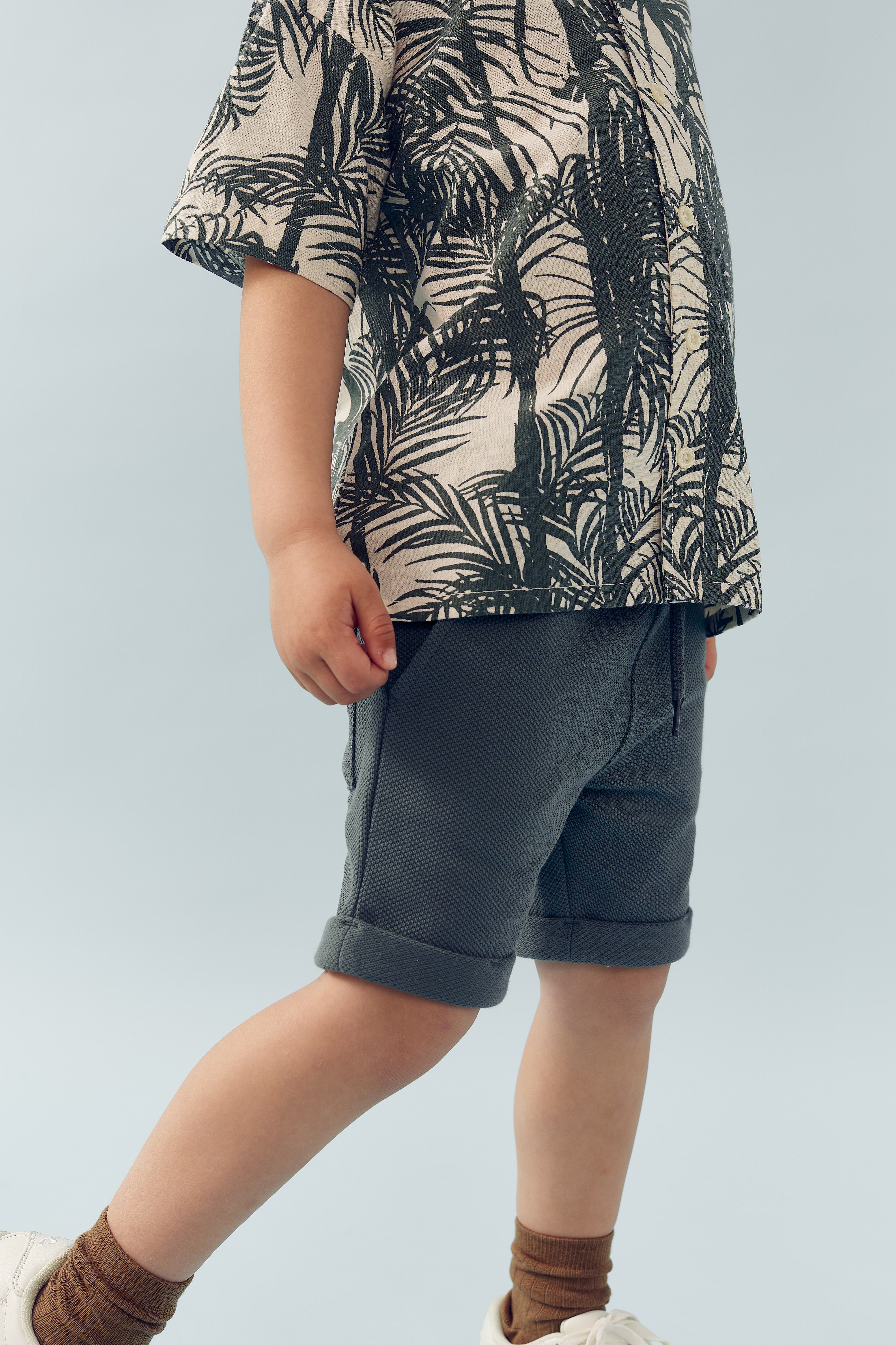 Child wearing a palm-patterned shirt and gray shorts stands in a neutral environment. Their feet are in white sneakers and brown socks. The background is a light blue shade.