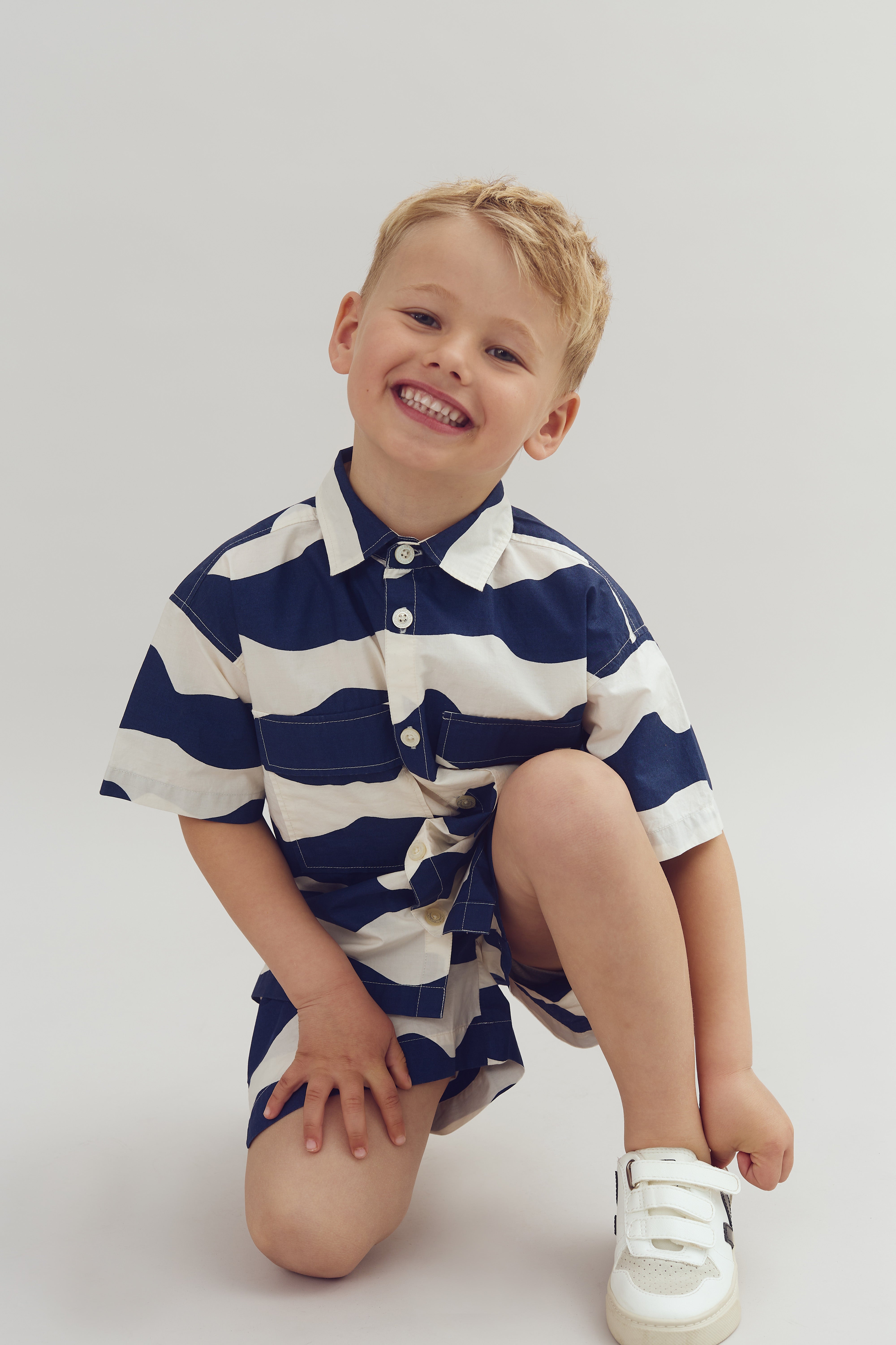 Child kneeling, smiling widely, wearing a navy and white striped shirt and shorts, with white shoes, against a plain background.