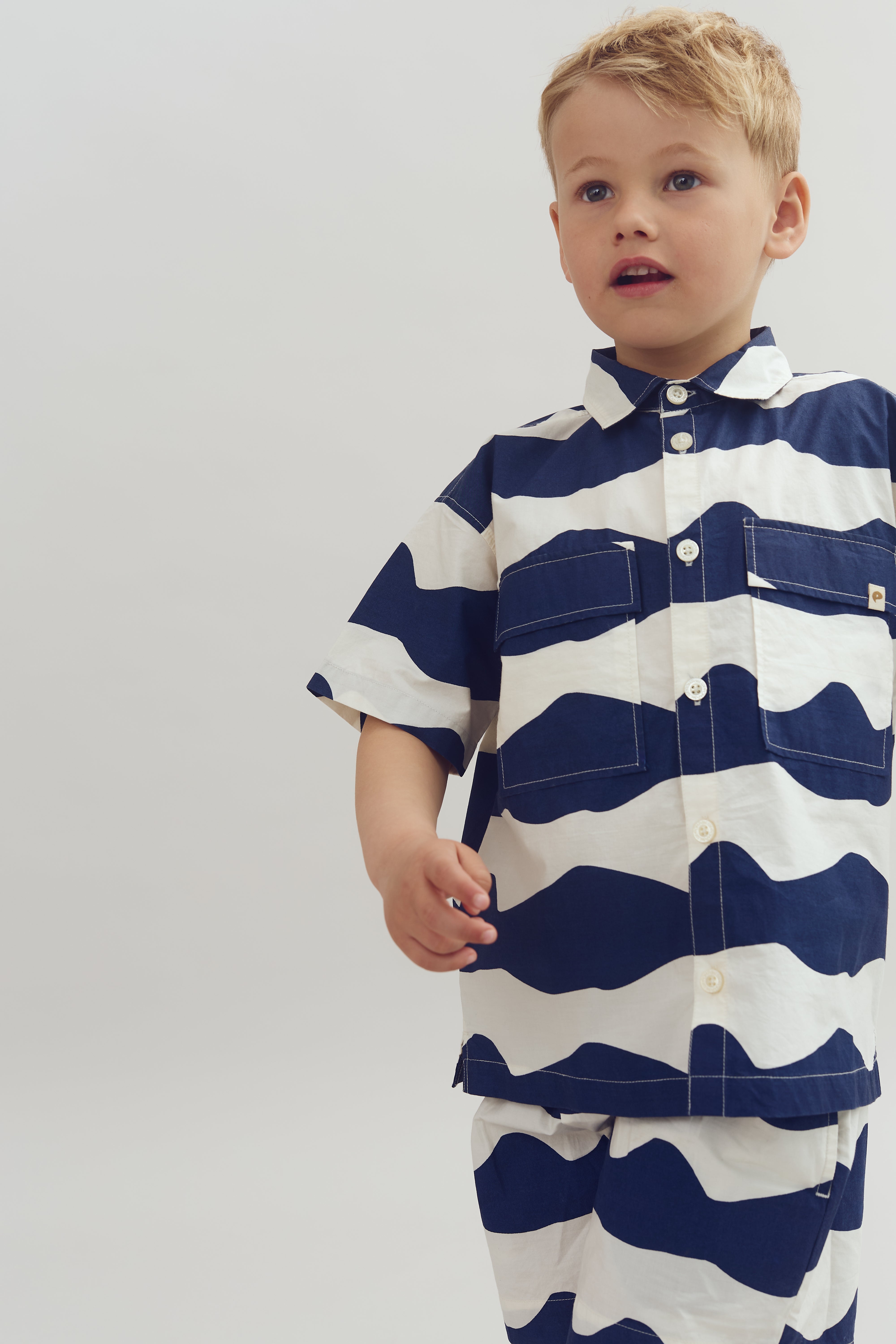 A child stands wearing a blue and white wavy patterned shirt and matching shorts, set against a plain light gray background.