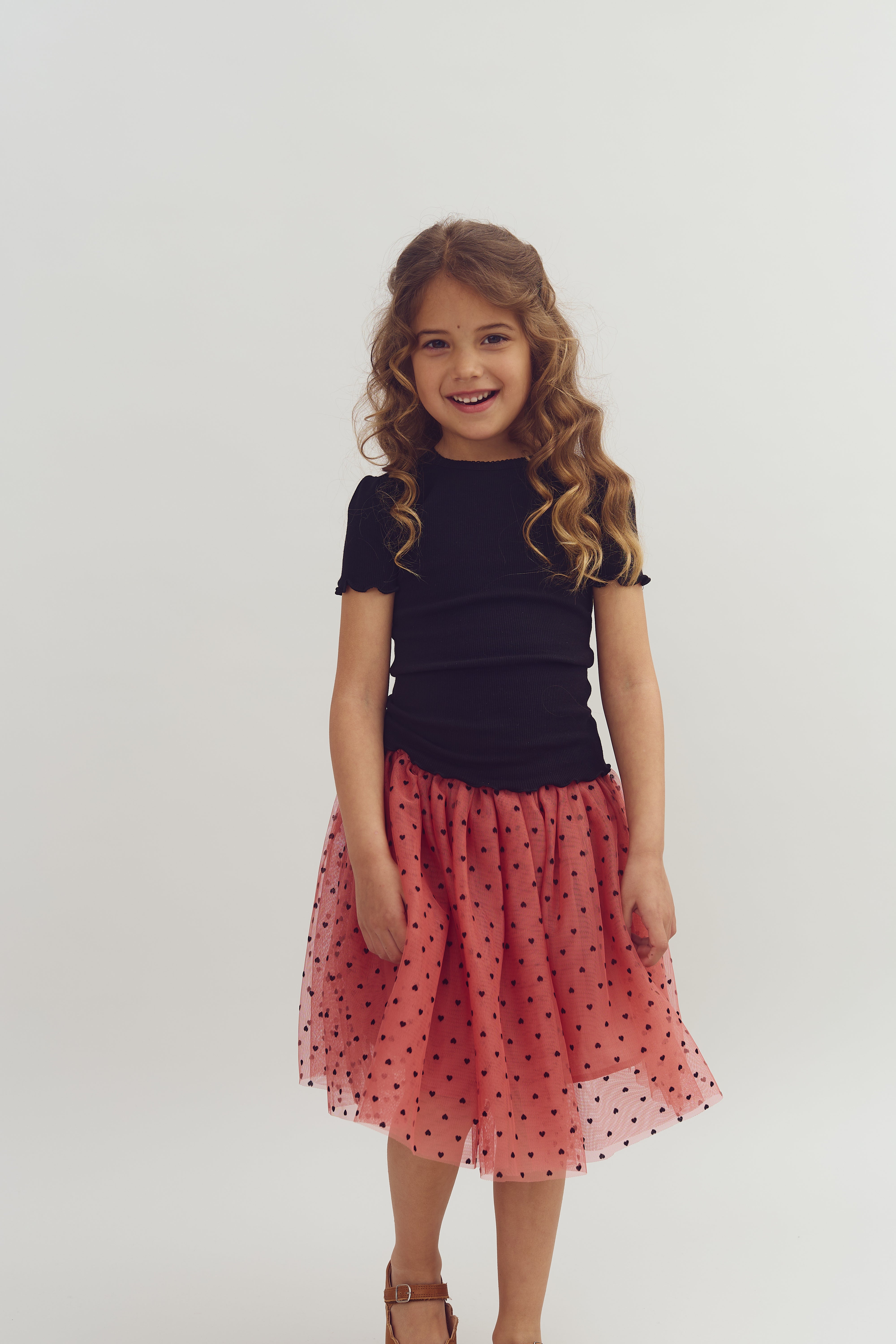 A young girl with curly hair smiles, wearing a black top and a pink polka dot skirt, in front of a plain white background.